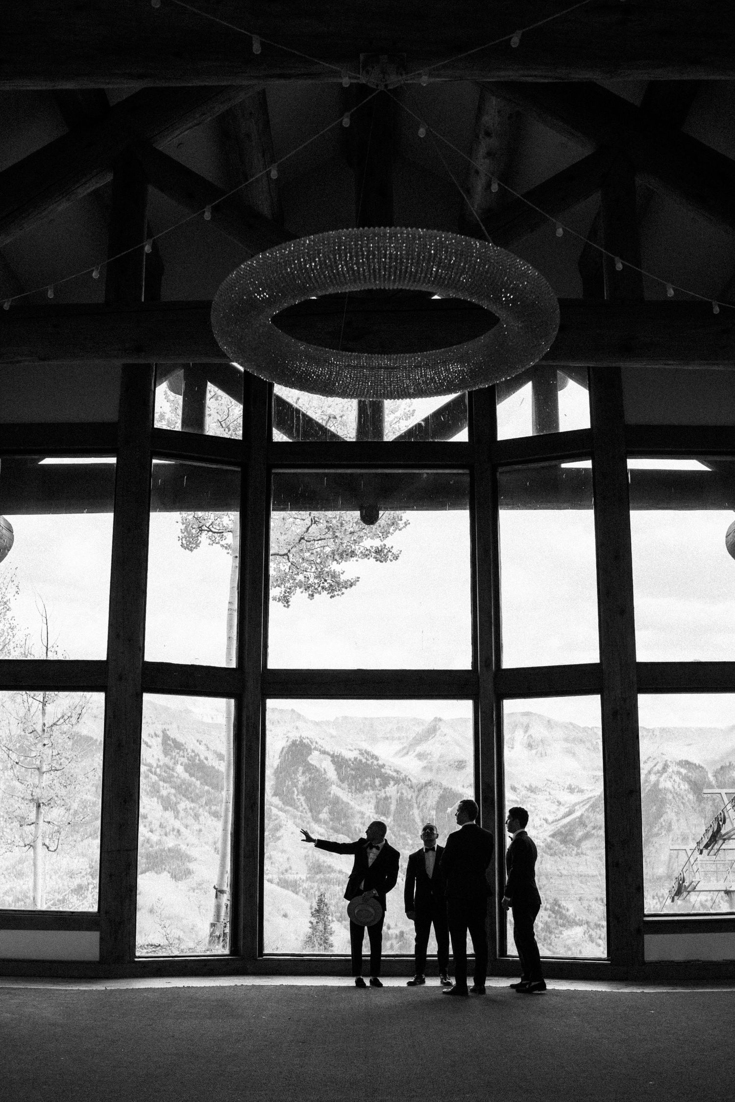 Groomsmen silhouetted against floor-to-ceiling windows with stunning mountain views in elegant wedding venue.