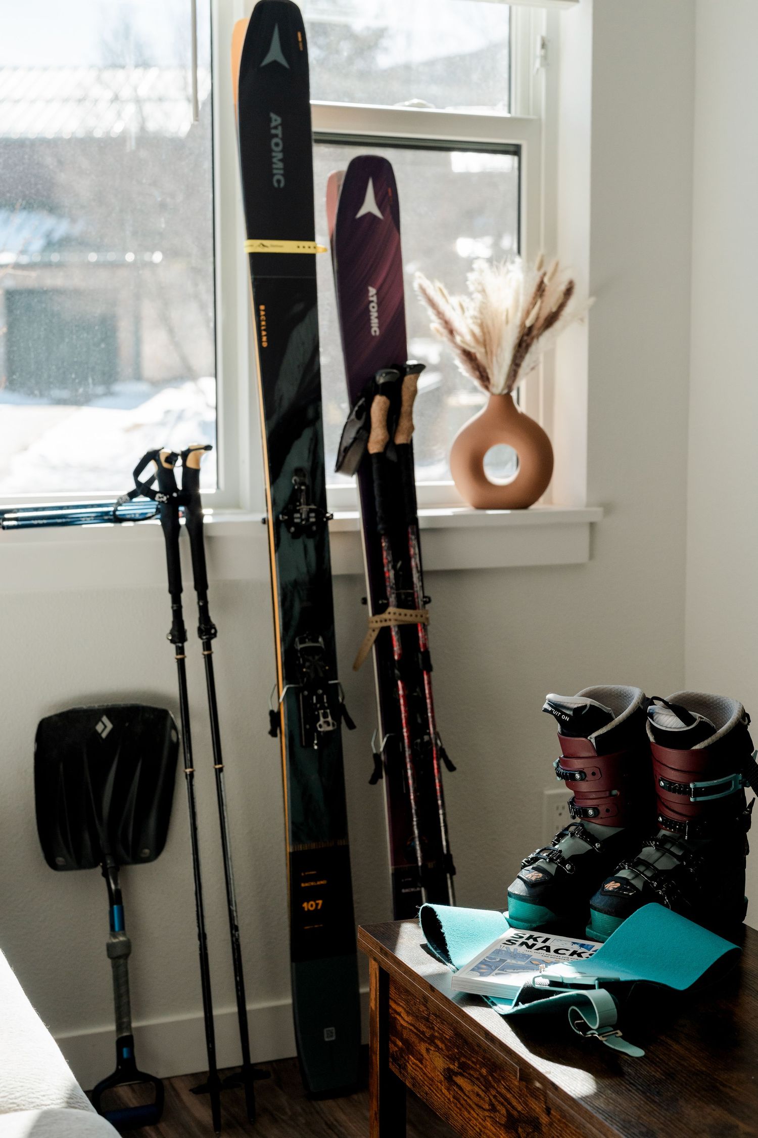 Atomic skis, poles, ski boots, and backcountry gear arranged near a window in a cozy home.