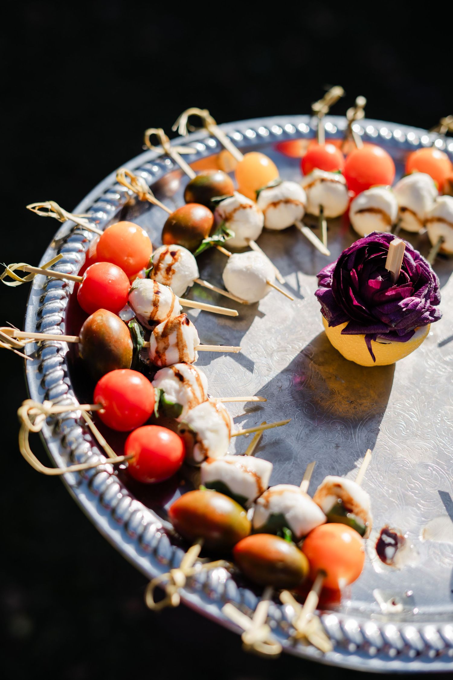 Colorful caprese and olive appetizer skewers arranged on silver tray with decorative purple flower garnish.