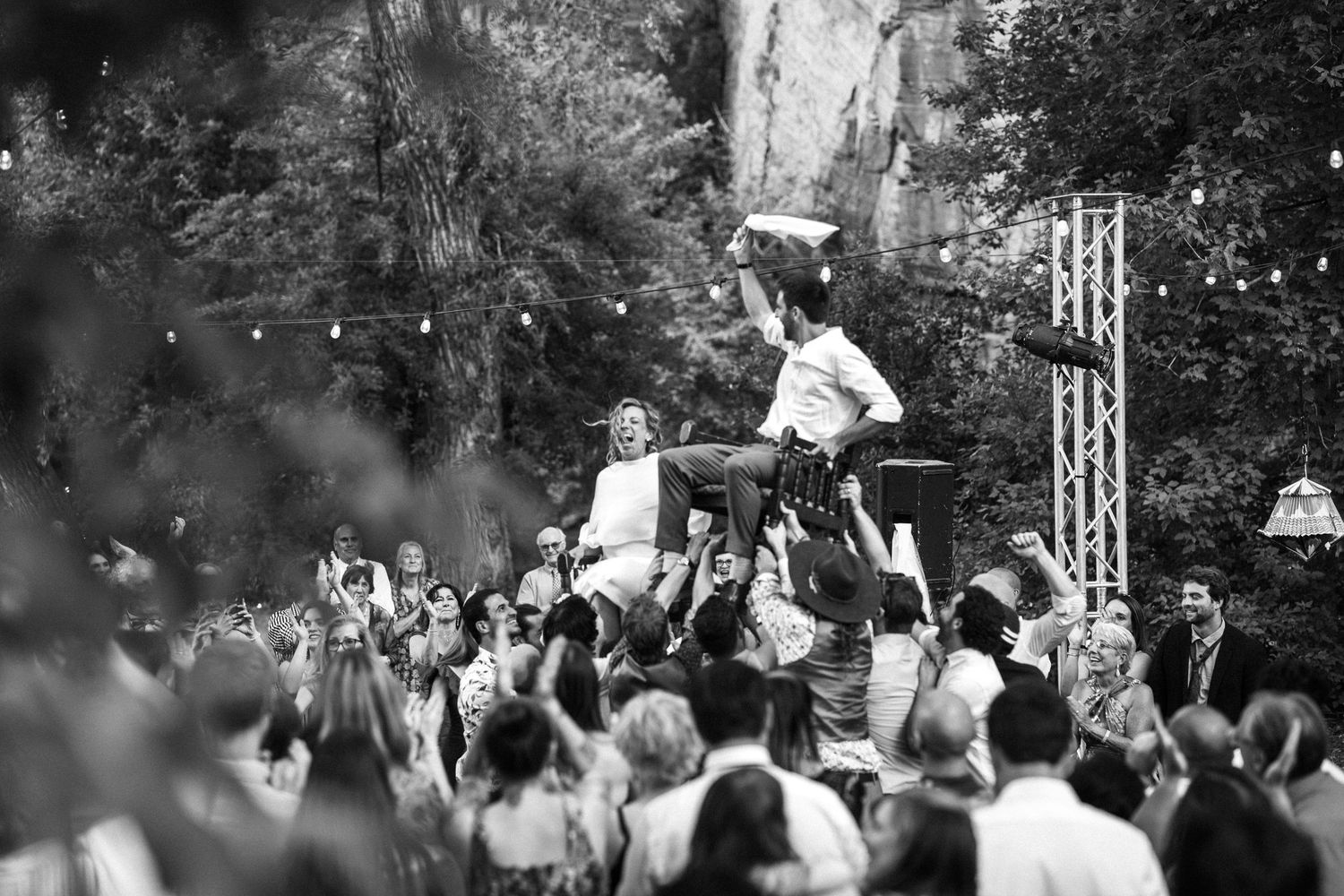 Black and white photo of wedding guests lifting couple in chairs at outdoor reception.