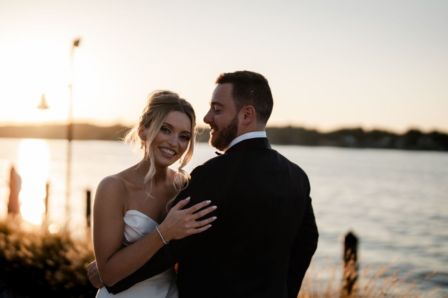 Bride smiling warmly as groom holds her close near a golden sunset waterfront at their wedding.