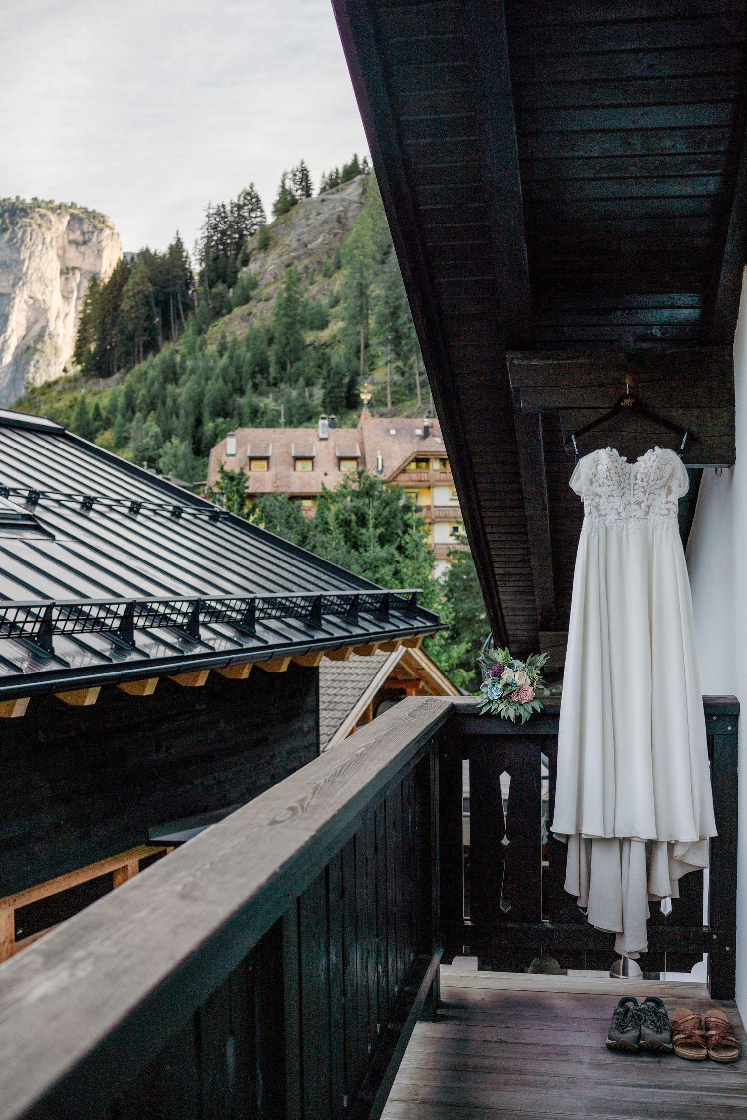 White lace wedding dress hangs on balcony railing with alpine mountain village backdrop in Switzerland.