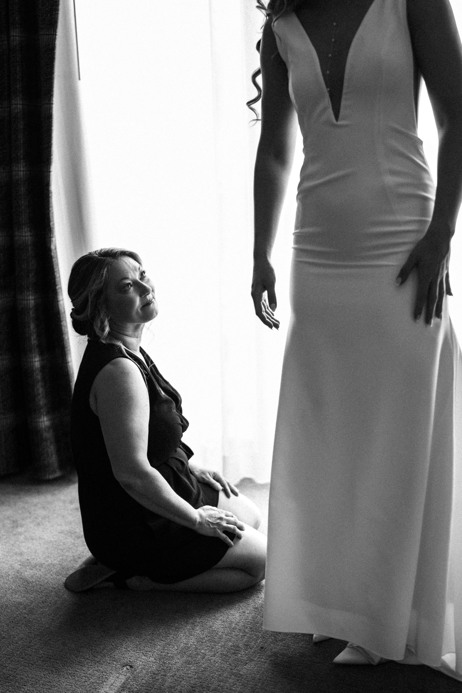 Black and white photo of woman kneeling before bride in wedding gown near a bright window.