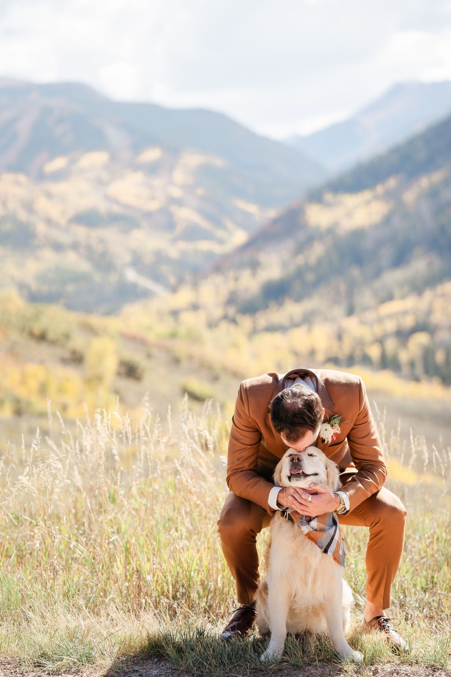 A groom in a brown suit tenderly embraces his golden retriever dog against a scenic mountain backdrop.