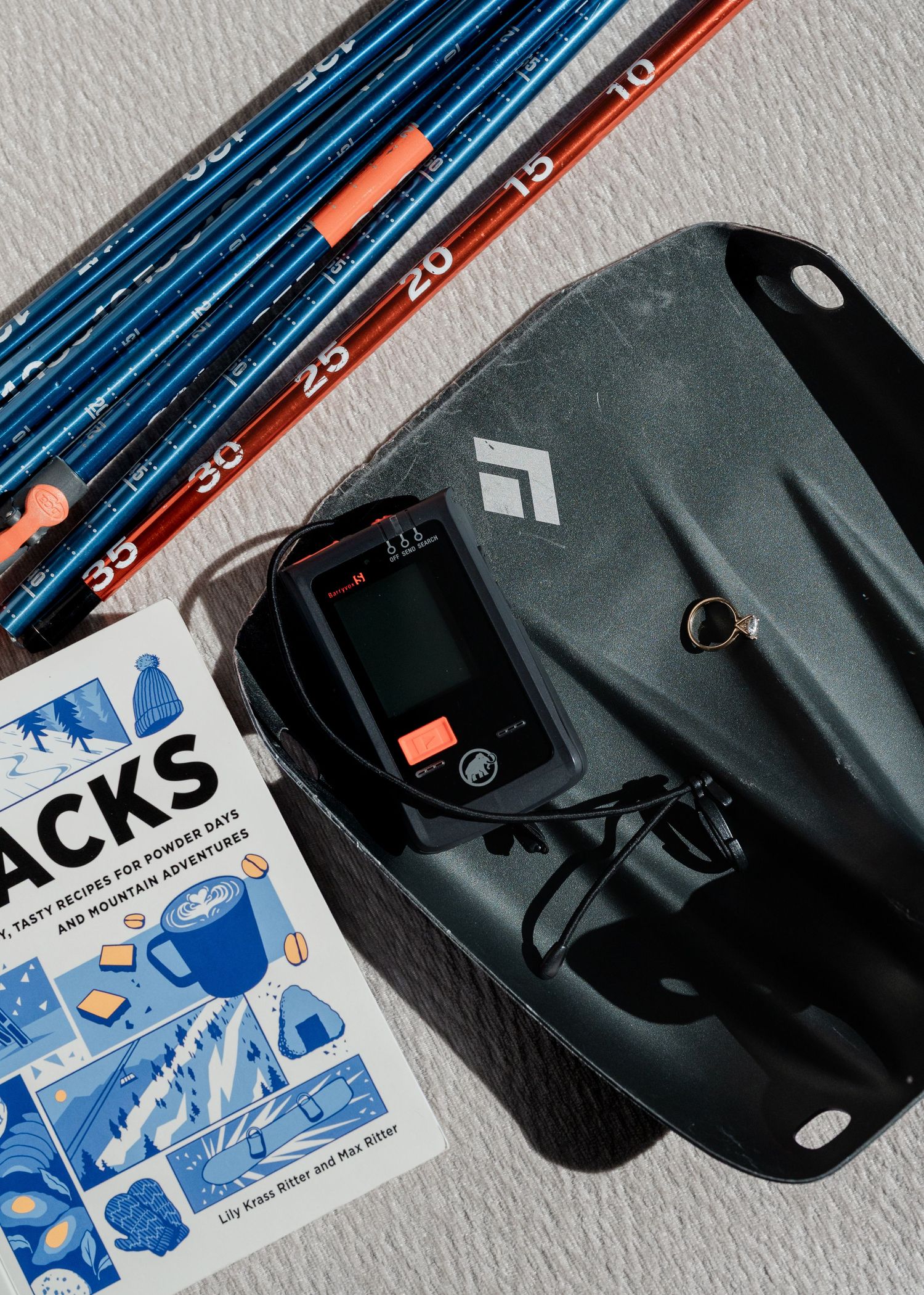 Avalanche safety gear including probes, beacon, and Black Diamond shovel with a mountain snacks cookbook.