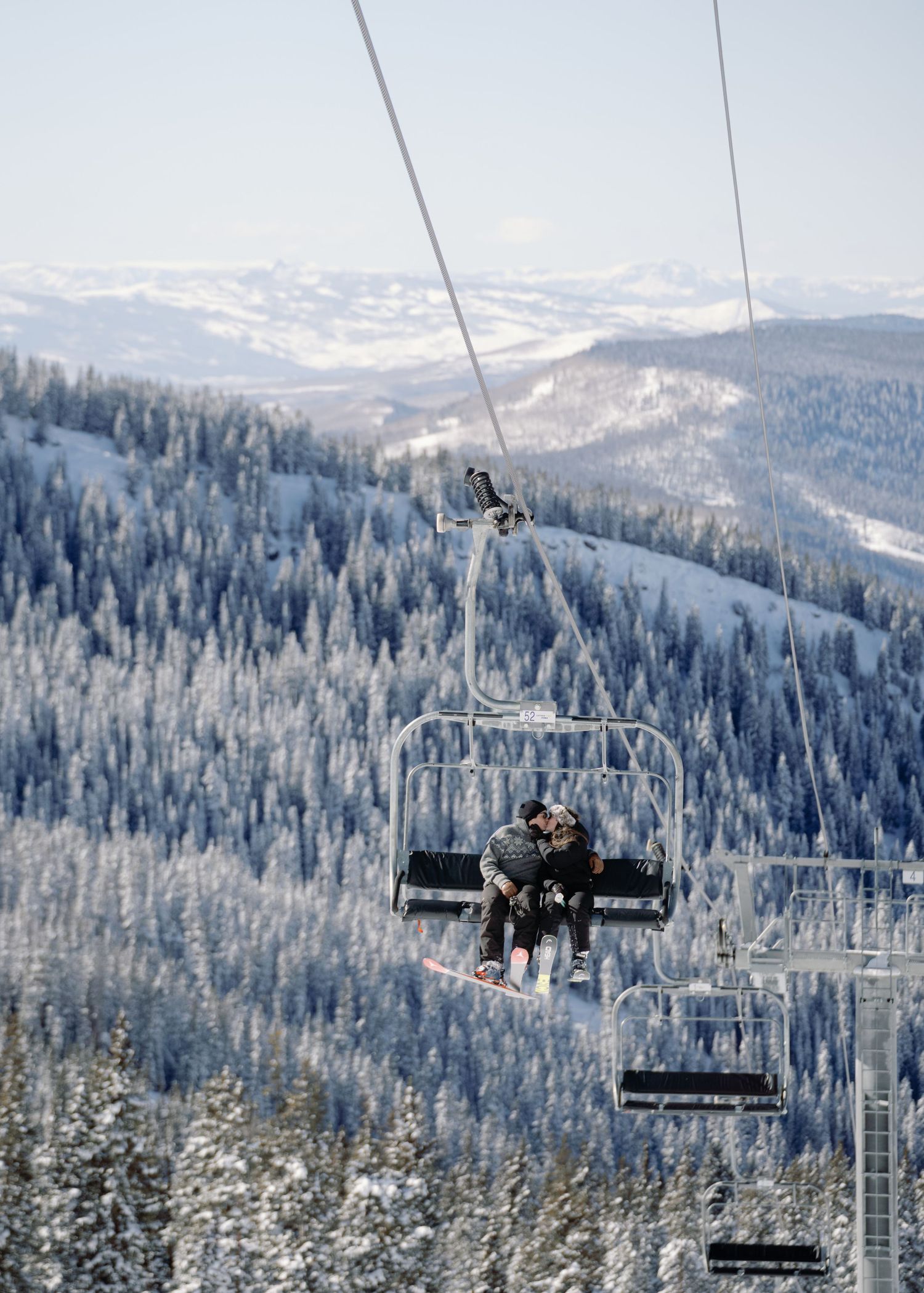 Two people riding a ski chairlift above a vast snow-covered mountain forest landscape.