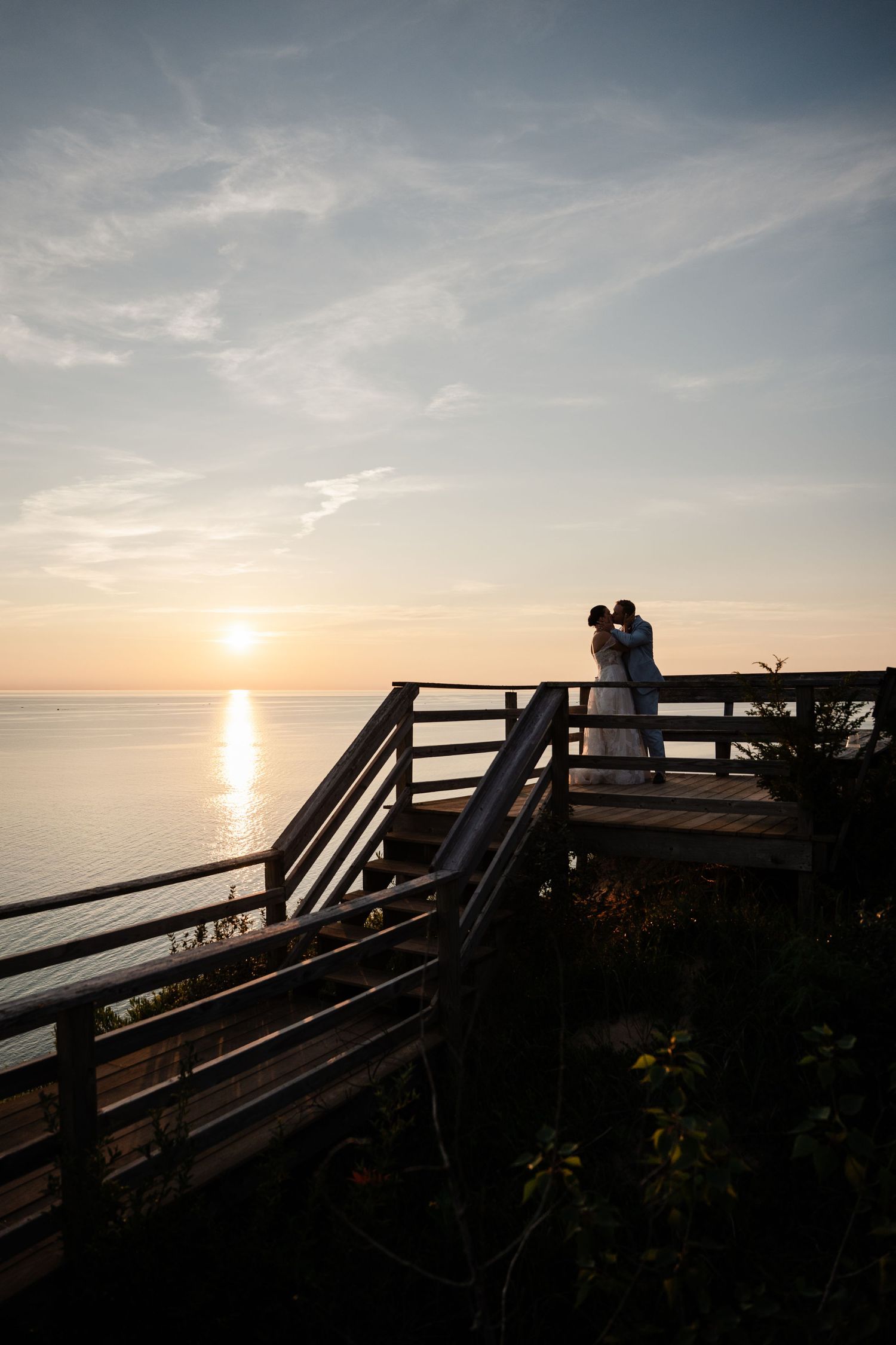 Newlyweds sharing a romantic kiss on a wooden overlook deck at a breathtaking ocean sunset.