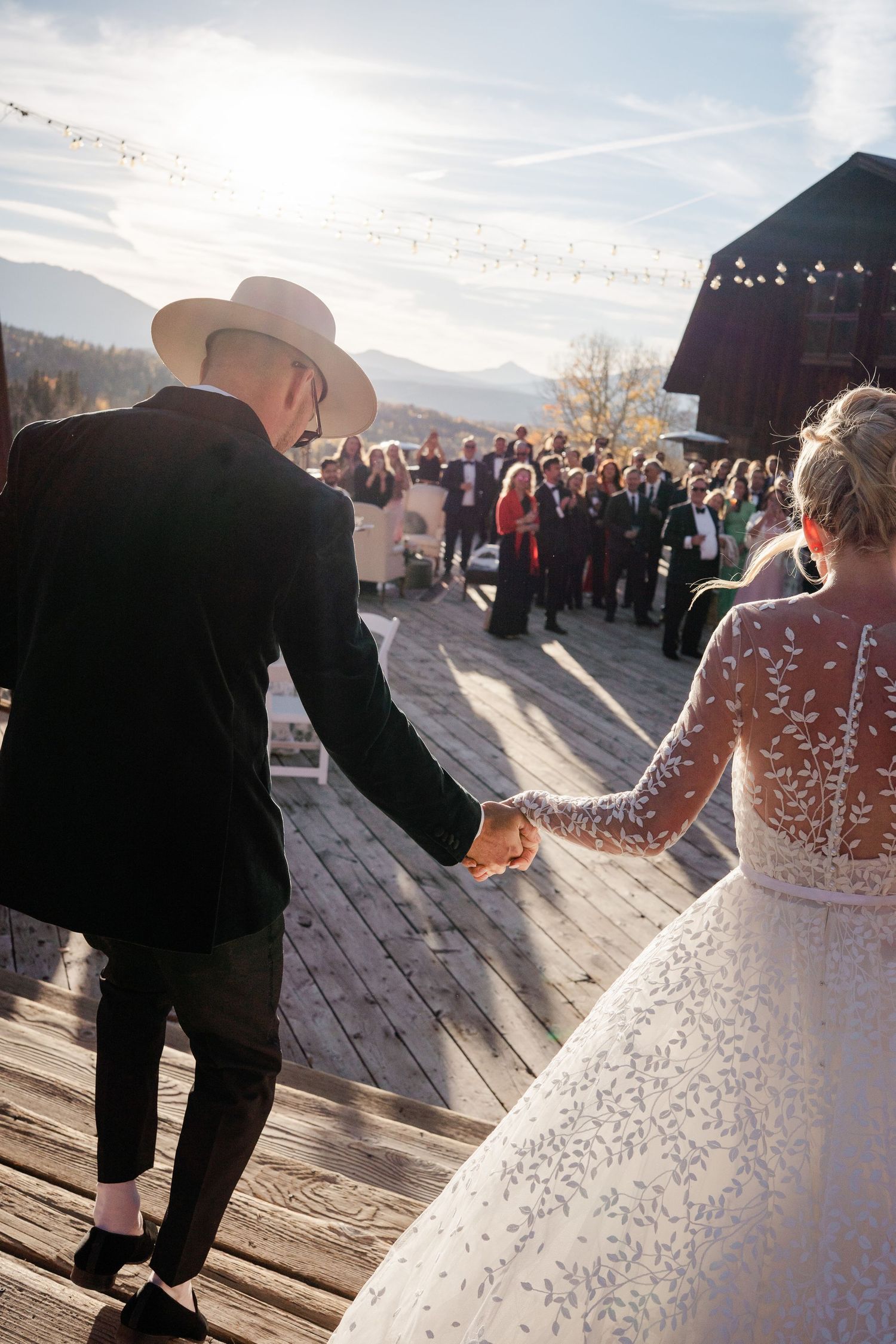 Bride and groom in cowboy hat hold hands walking toward wedding guests at mountain venue.