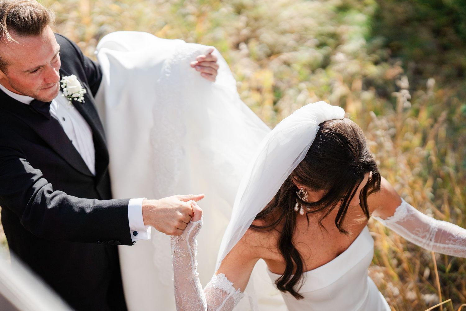 Groom in black tuxedo lifting bride's white veil and dress while walking through autumn field.