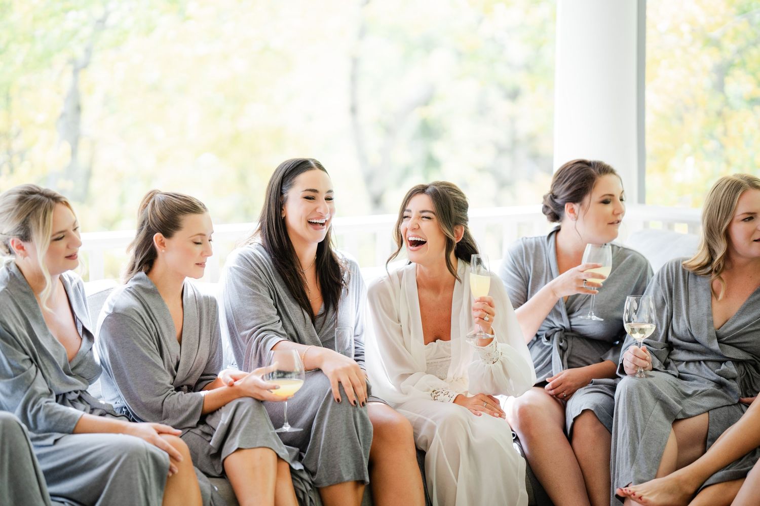 Bride and bridesmaids laughing together in matching grey robes while holding champagne glasses during wedding preparations.