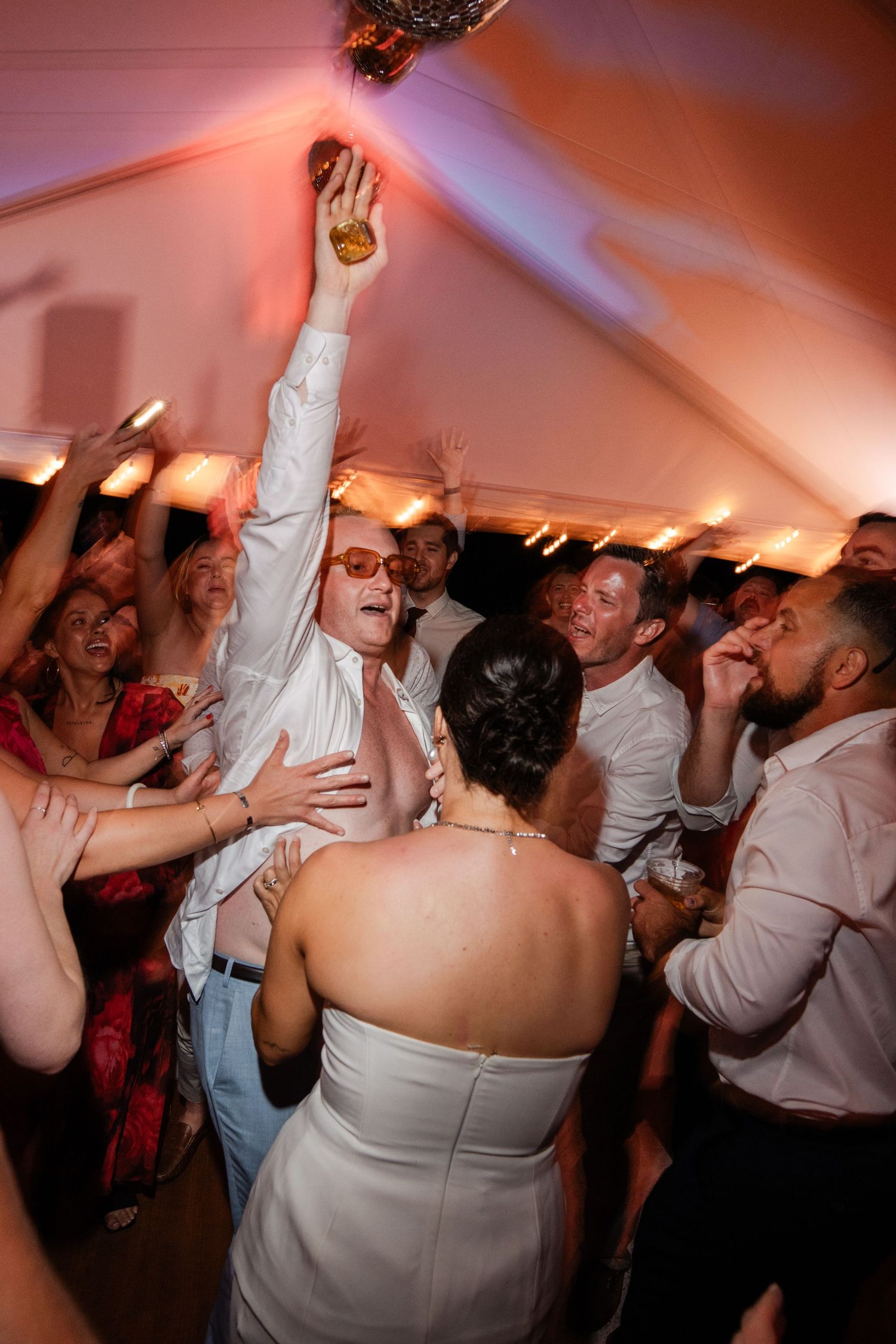 Energetic wedding guests dance wildly under disco balls and warm pink lighting in a tent.