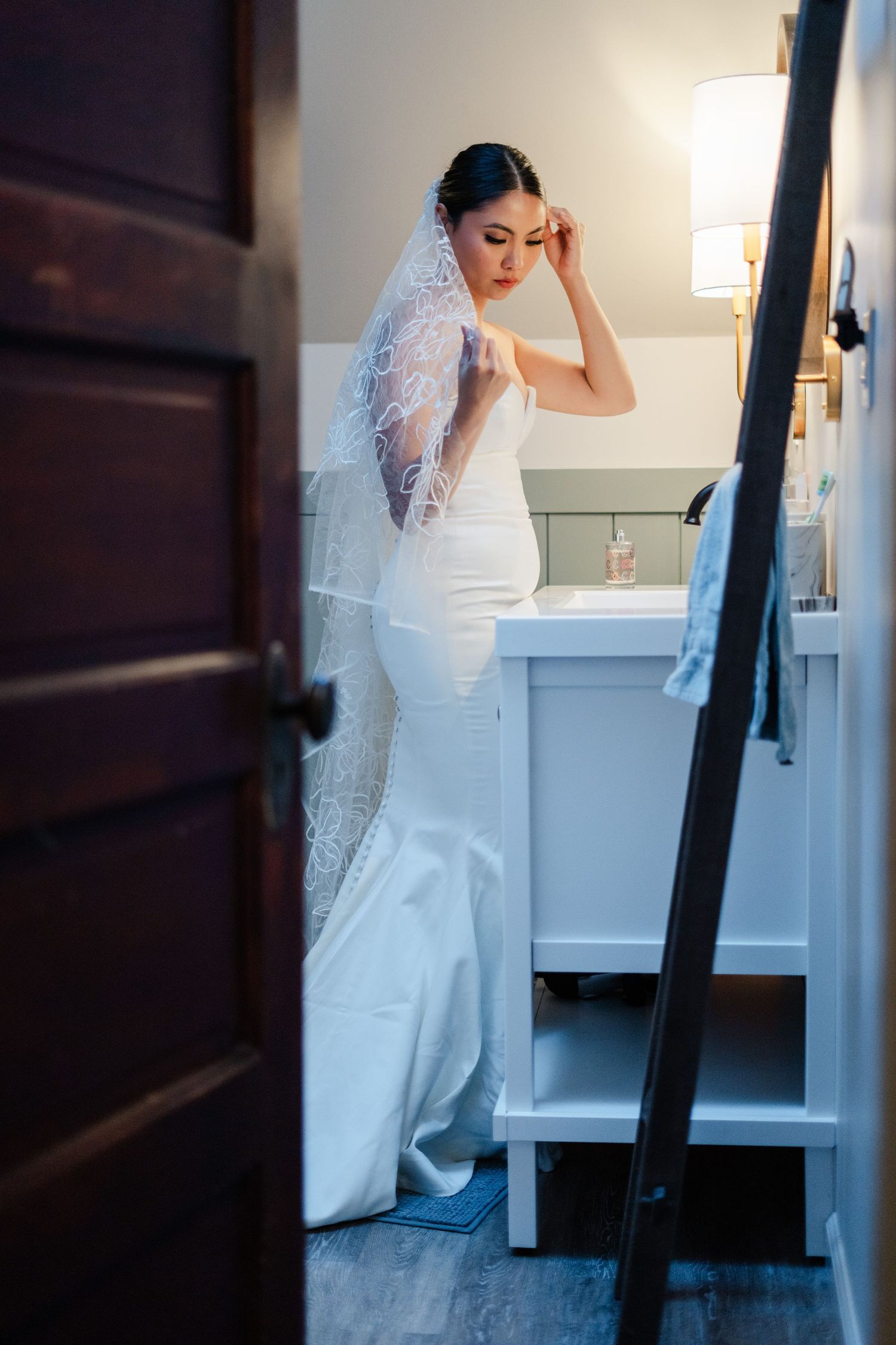Bride in elegant white mermaid gown and lace veil preparing in bathroom mirror before wedding ceremony.