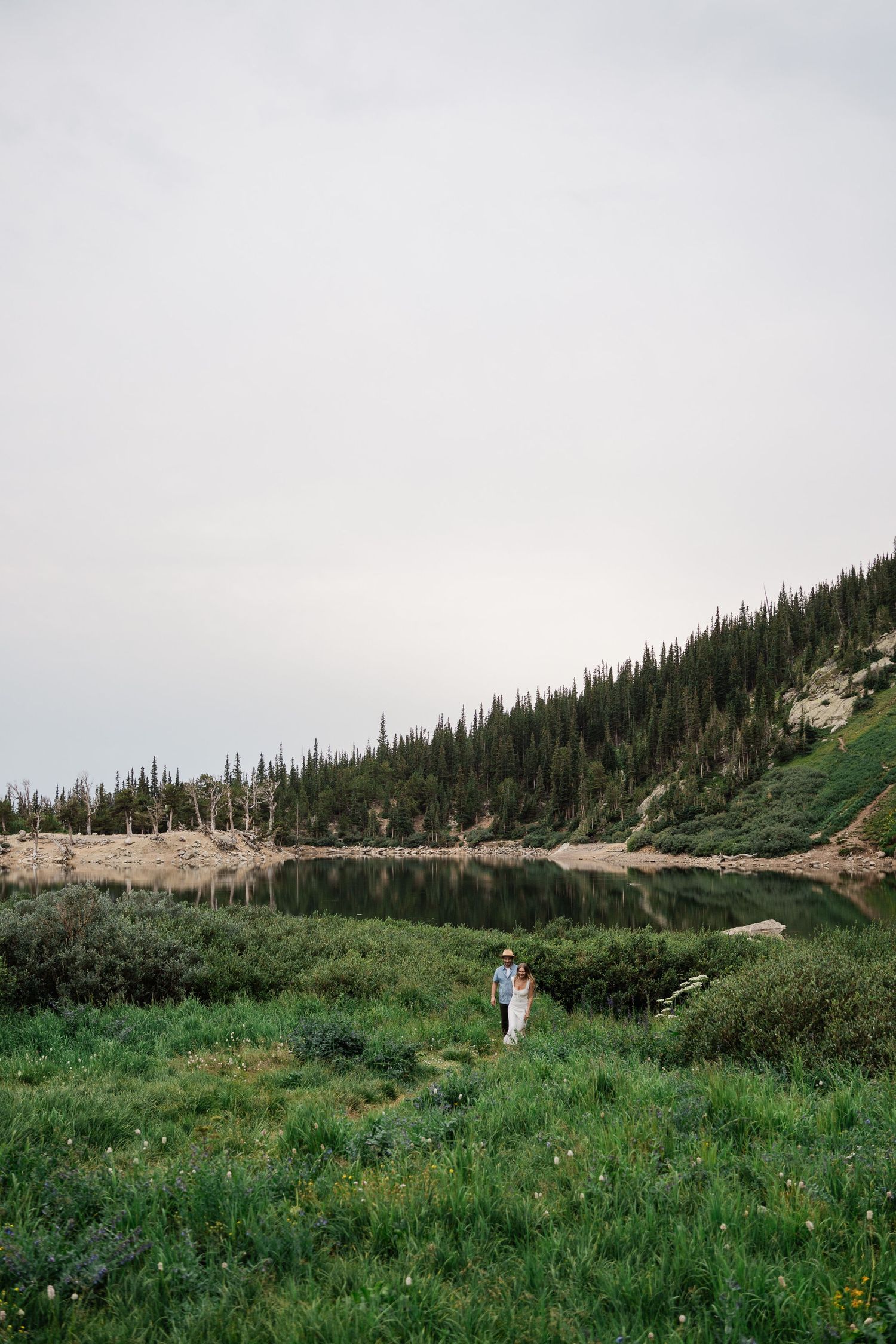 Couple embracing in a lush green meadow beside a serene mountain lake with pine tree reflections.