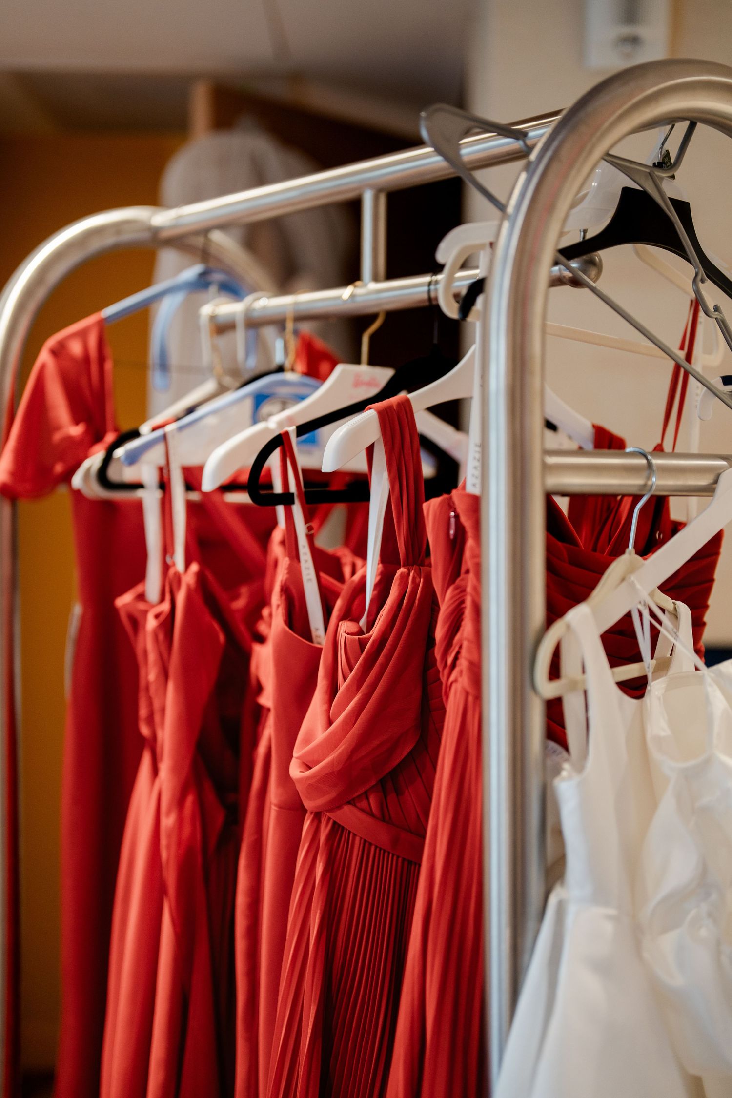 Red bridesmaid dresses hanging on silver clothing rack with white garment bags in bridal suite.