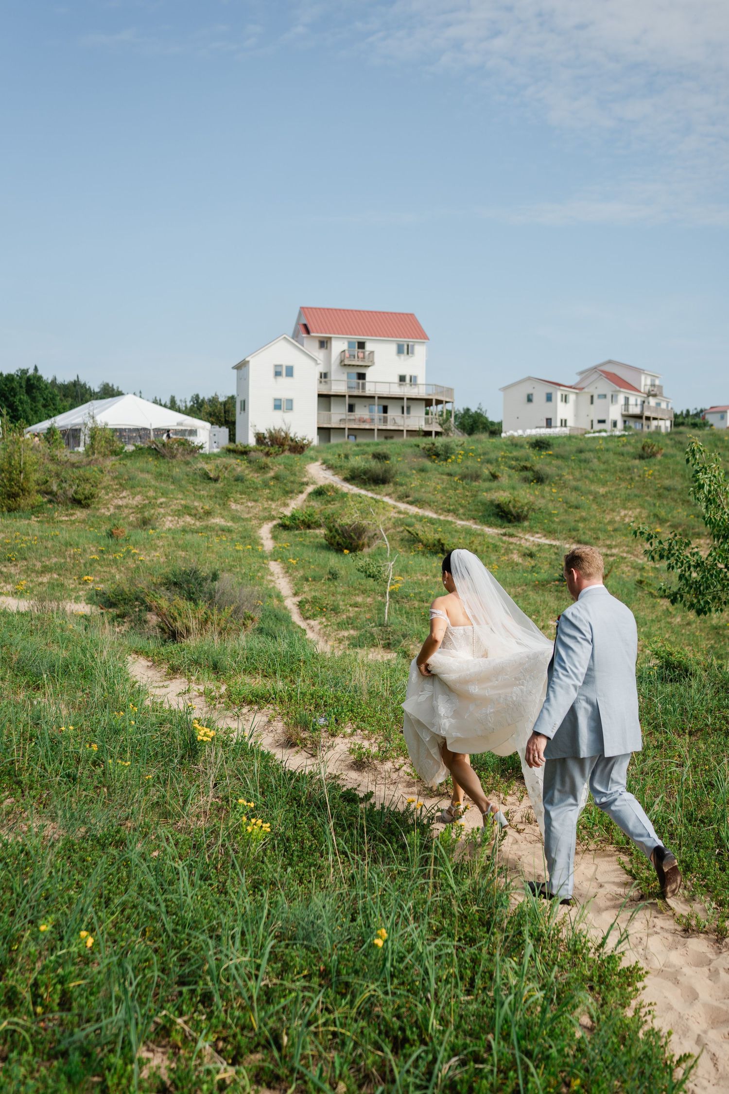Bride and groom walk sandy path toward white multi-story coastal wedding venue on a sunny day.