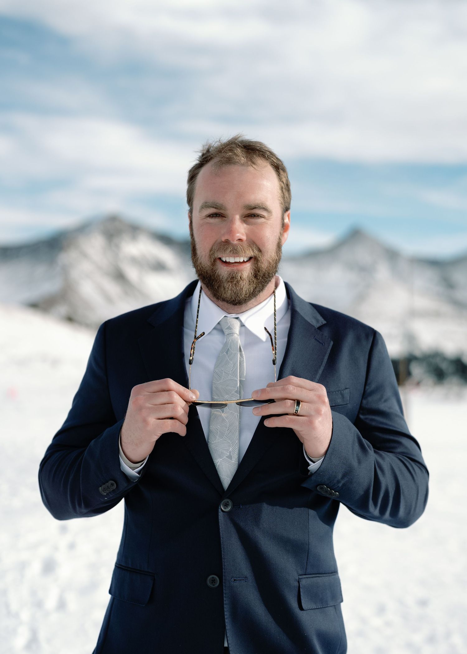 Bearded groom in navy suit smiles while holding sunglasses on a snowy mountain wedding day.