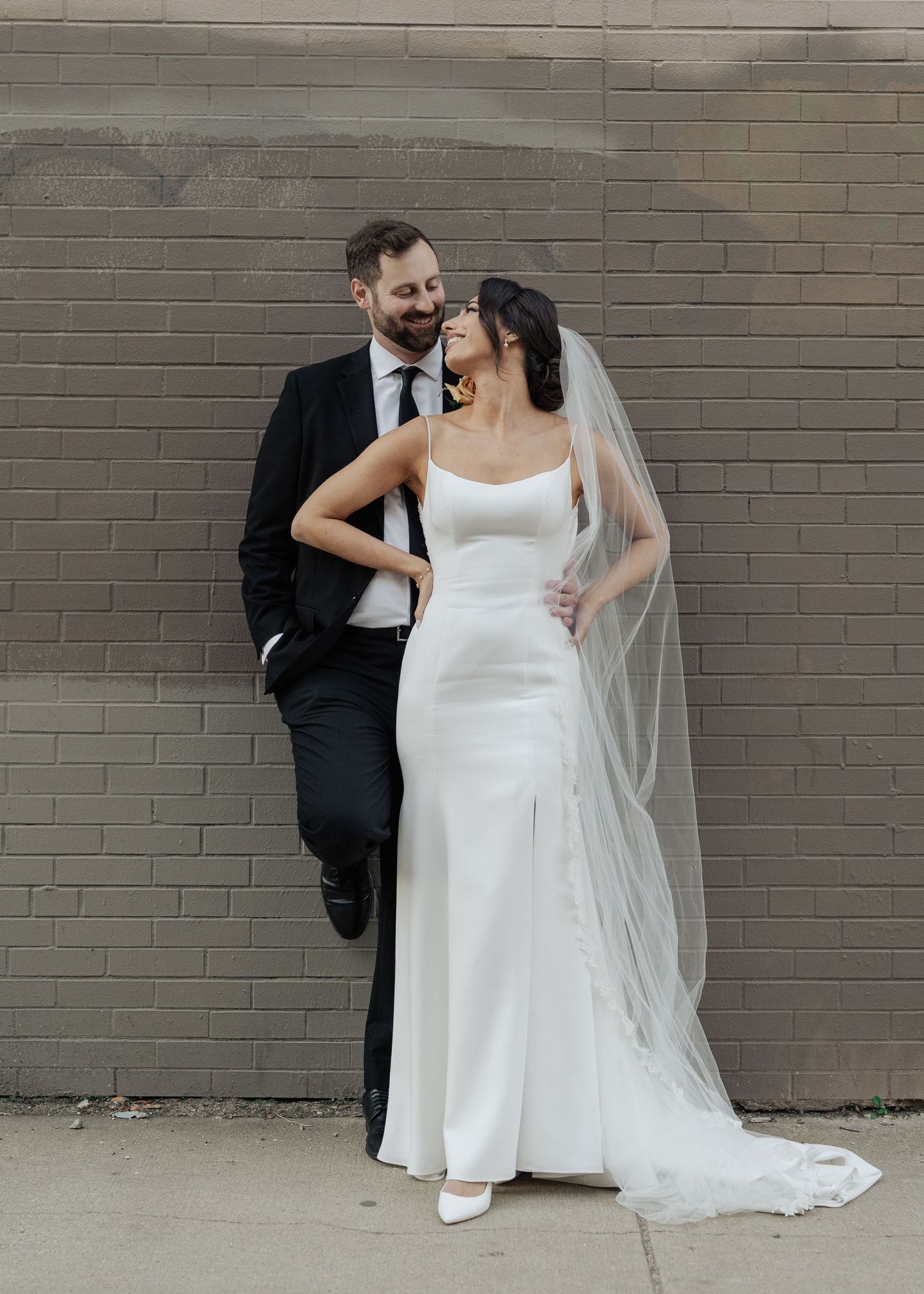 Stylish bride and groom sharing a laugh while posing against an urban brick wall.