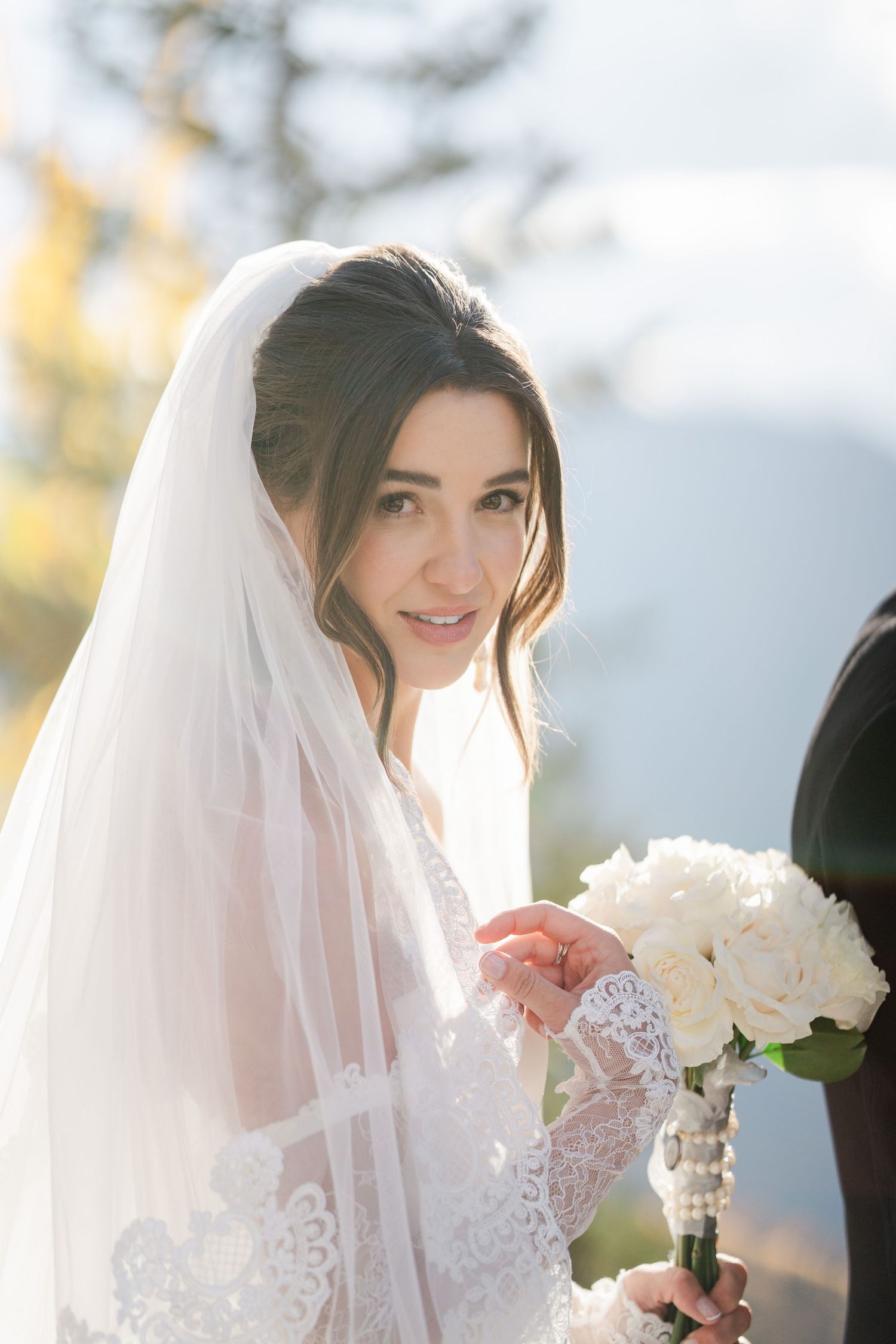 Beautiful bride in lace wedding gown and veil holds white rose bouquet with soft mountain light behind her.