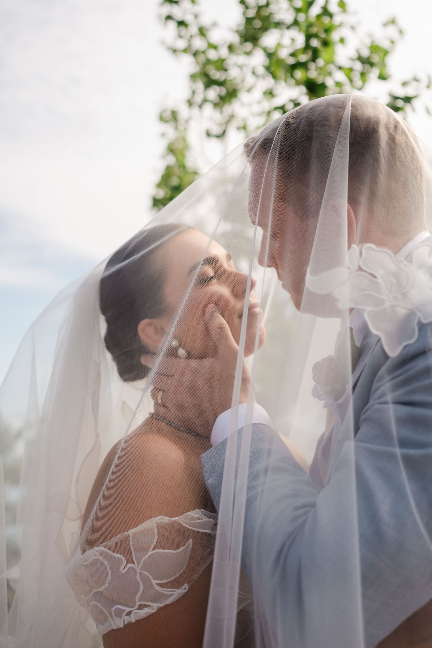 Bride and groom sharing intimate moment under flowing wedding veil outdoors.