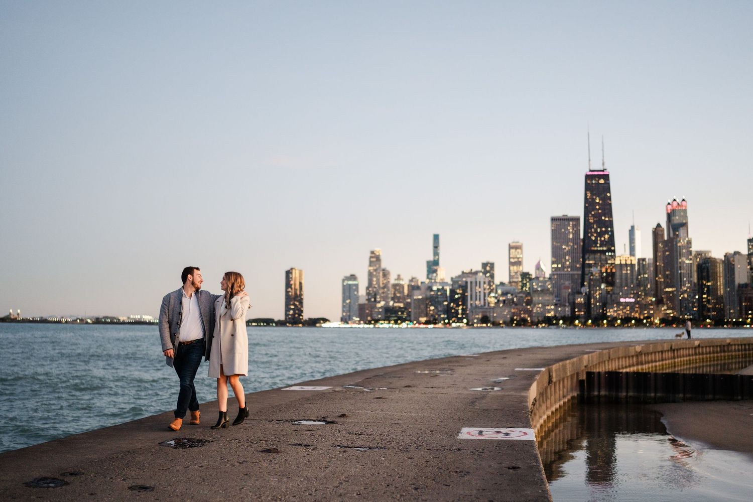 Engaged couple walking hand in hand along Chicago lakefront with stunning illuminated city skyline at dusk.