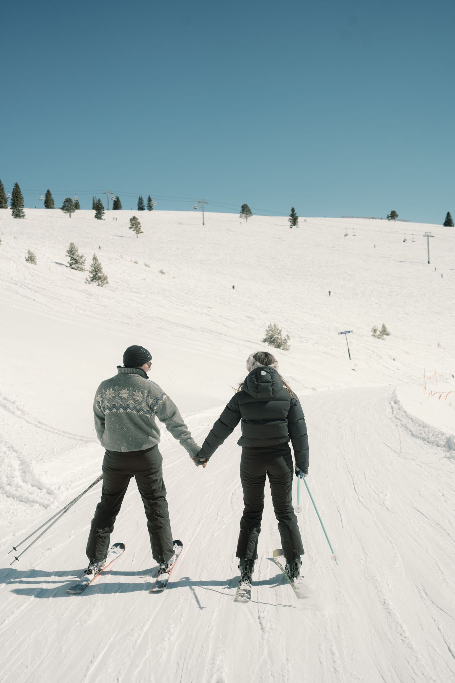 Couple holding hands while skiing down a snowy slope on a clear blue sky winter day.