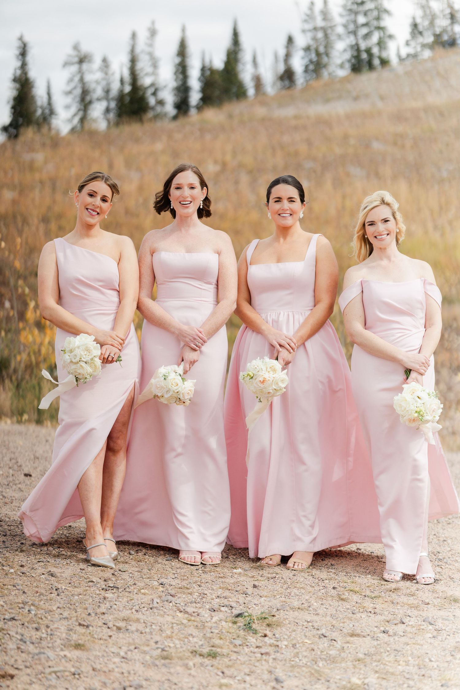Four bridesmaids in blush pink gowns holding white bouquets posing in front of autumn mountain landscape.