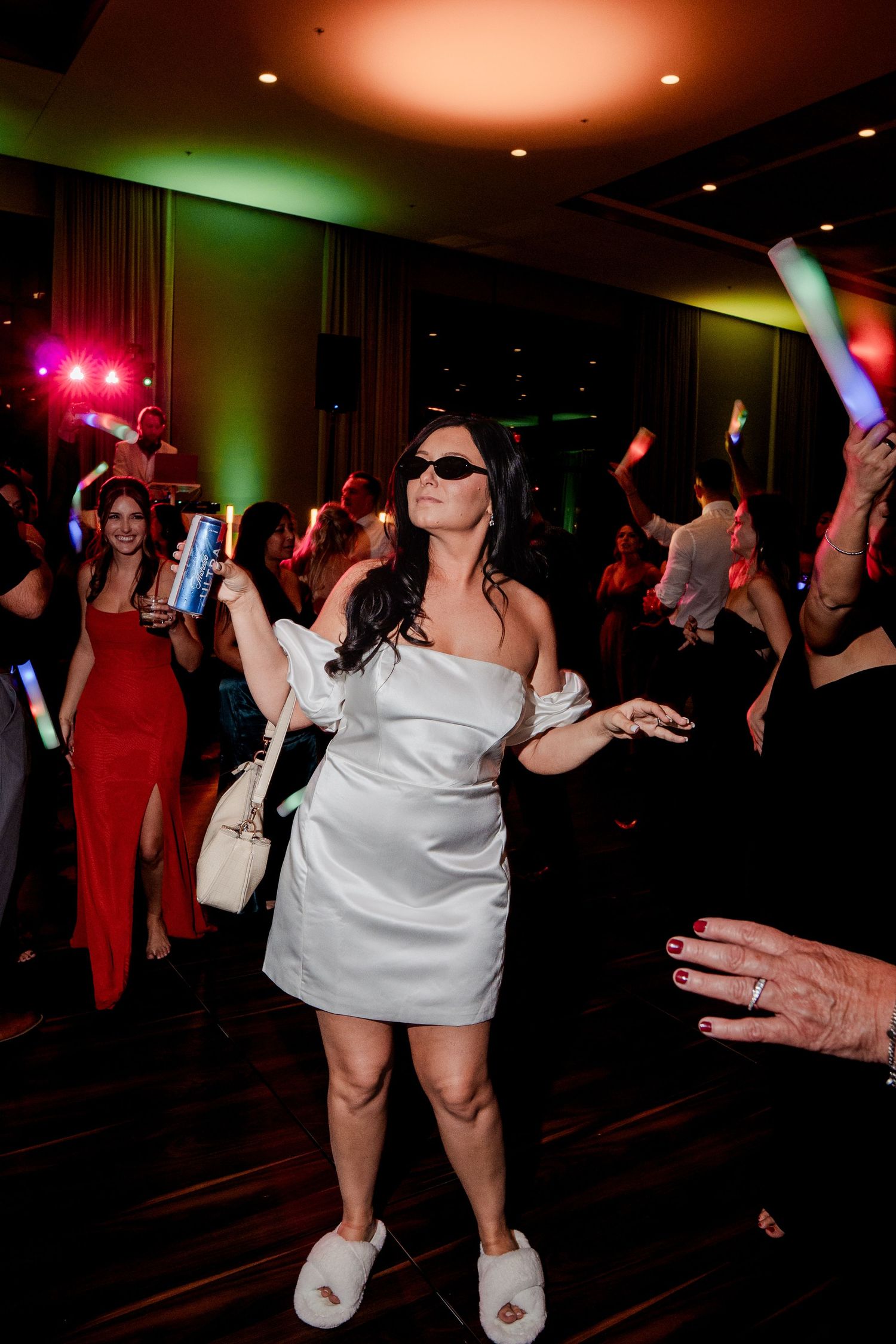 Bride in white mini dress and slippers wearing sunglasses dancing with guests at wedding reception.