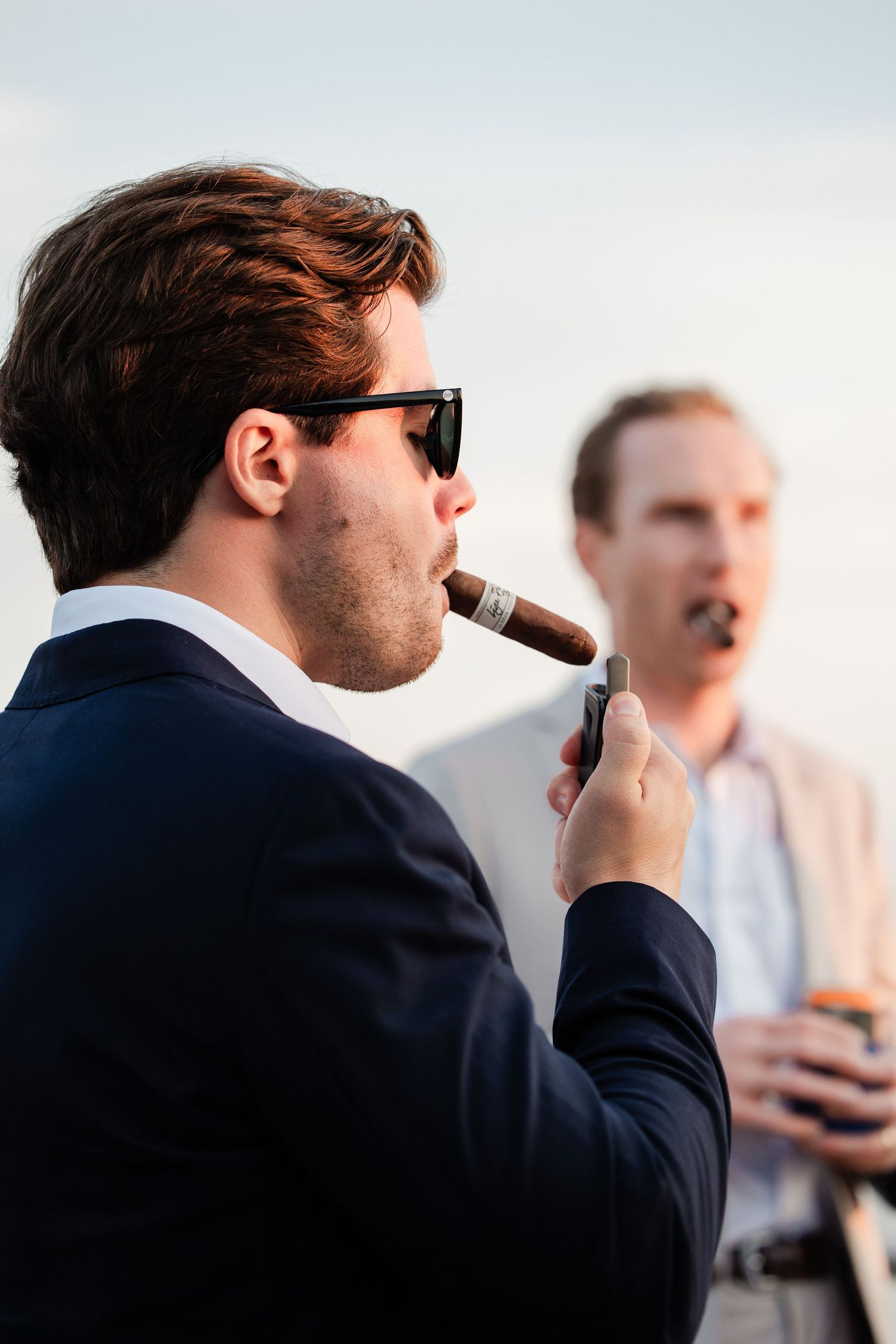 Two men in suits smoking cigars outdoors during a wedding celebration at sunset.
