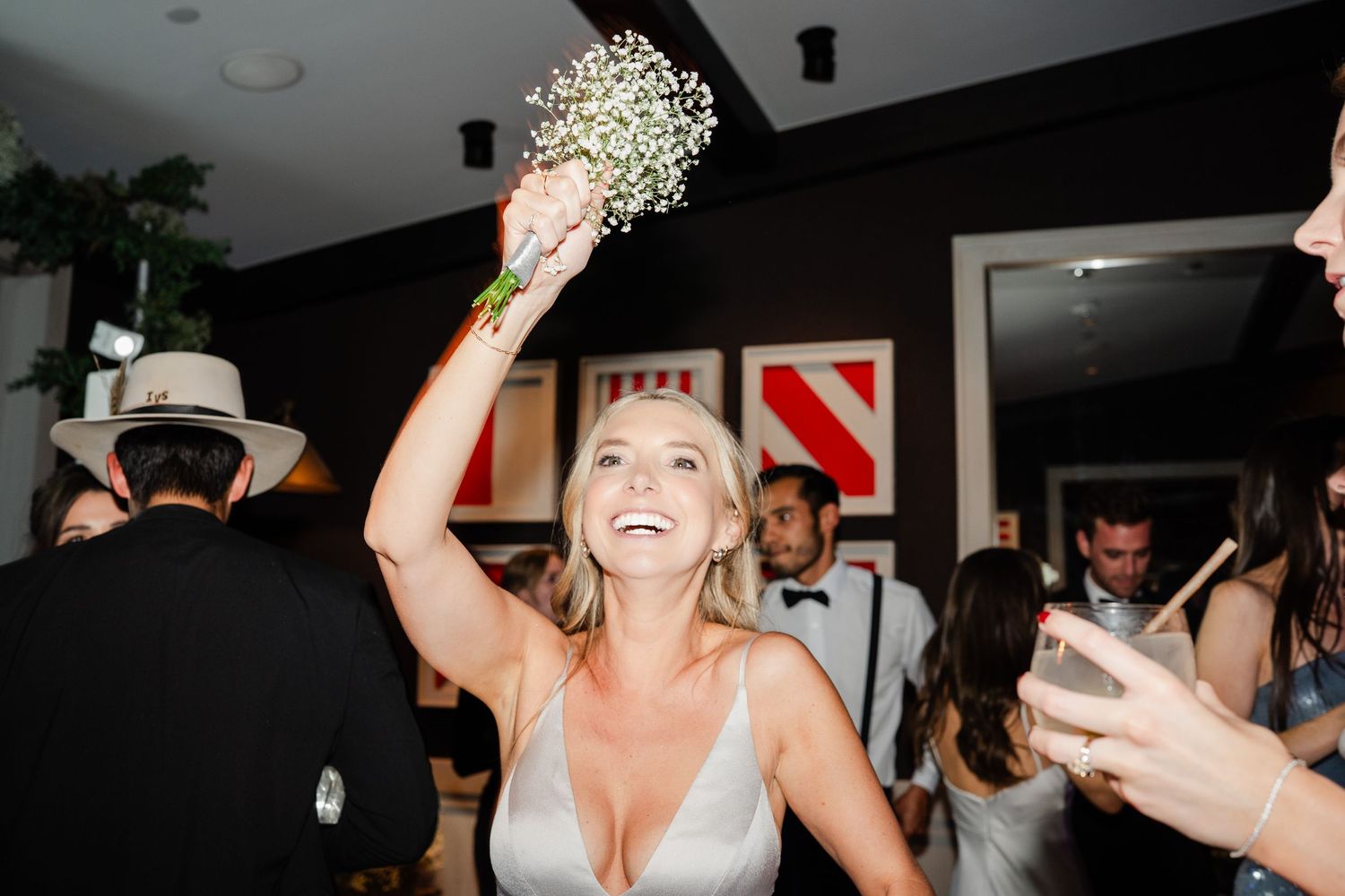 Smiling bridesmaid in silver dress holds bouquet overhead on the wedding reception dance floor.