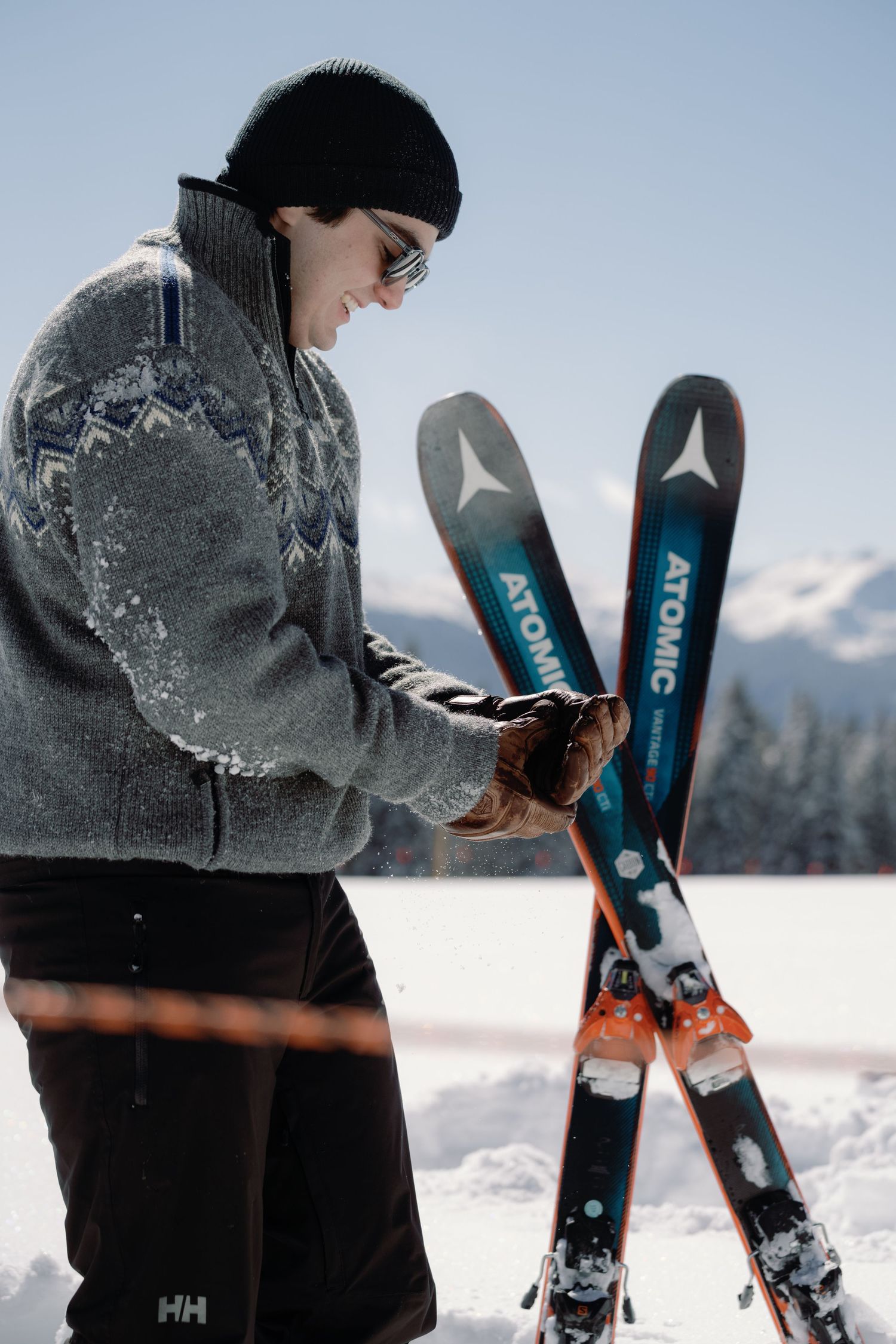 Smiling skier in winter hat holding Atomic skis on a sunny snowy mountain slope.