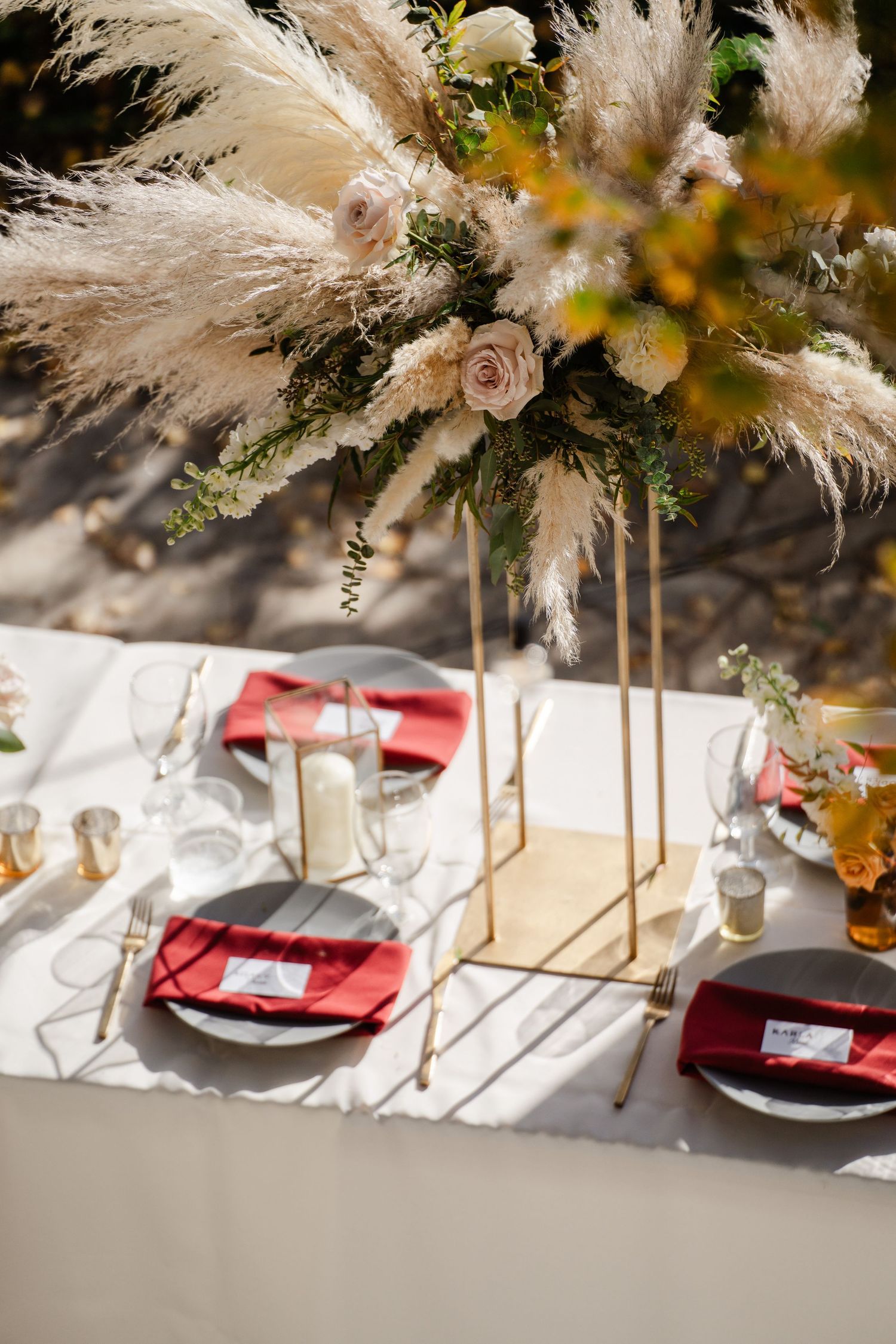 An elegant outdoor wedding tablescape with pampas grass centerpiece, red napkins, and gold accents.