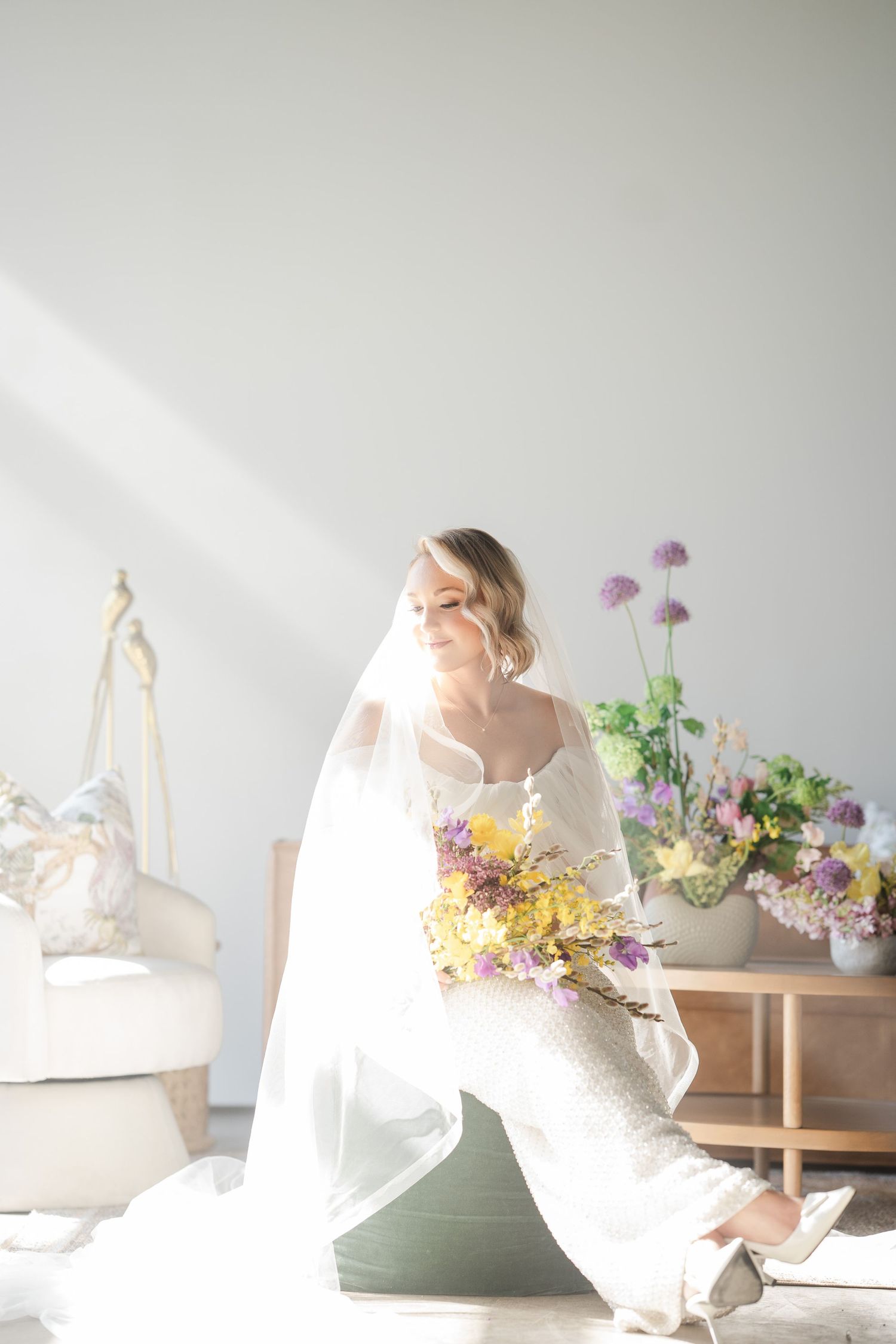 Radiant bride in a white gown and veil sits in sunlight holding a colorful wildflower bouquet indoors.