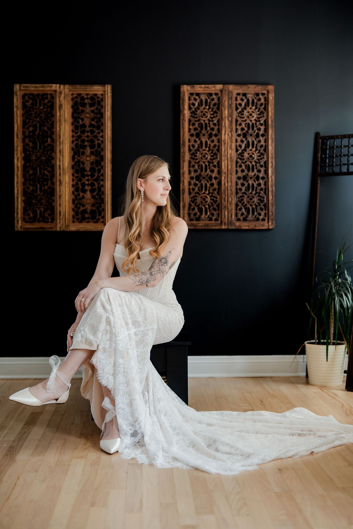 Bride in lace wedding gown seated elegantly against a dark wall with ornate wooden panels.