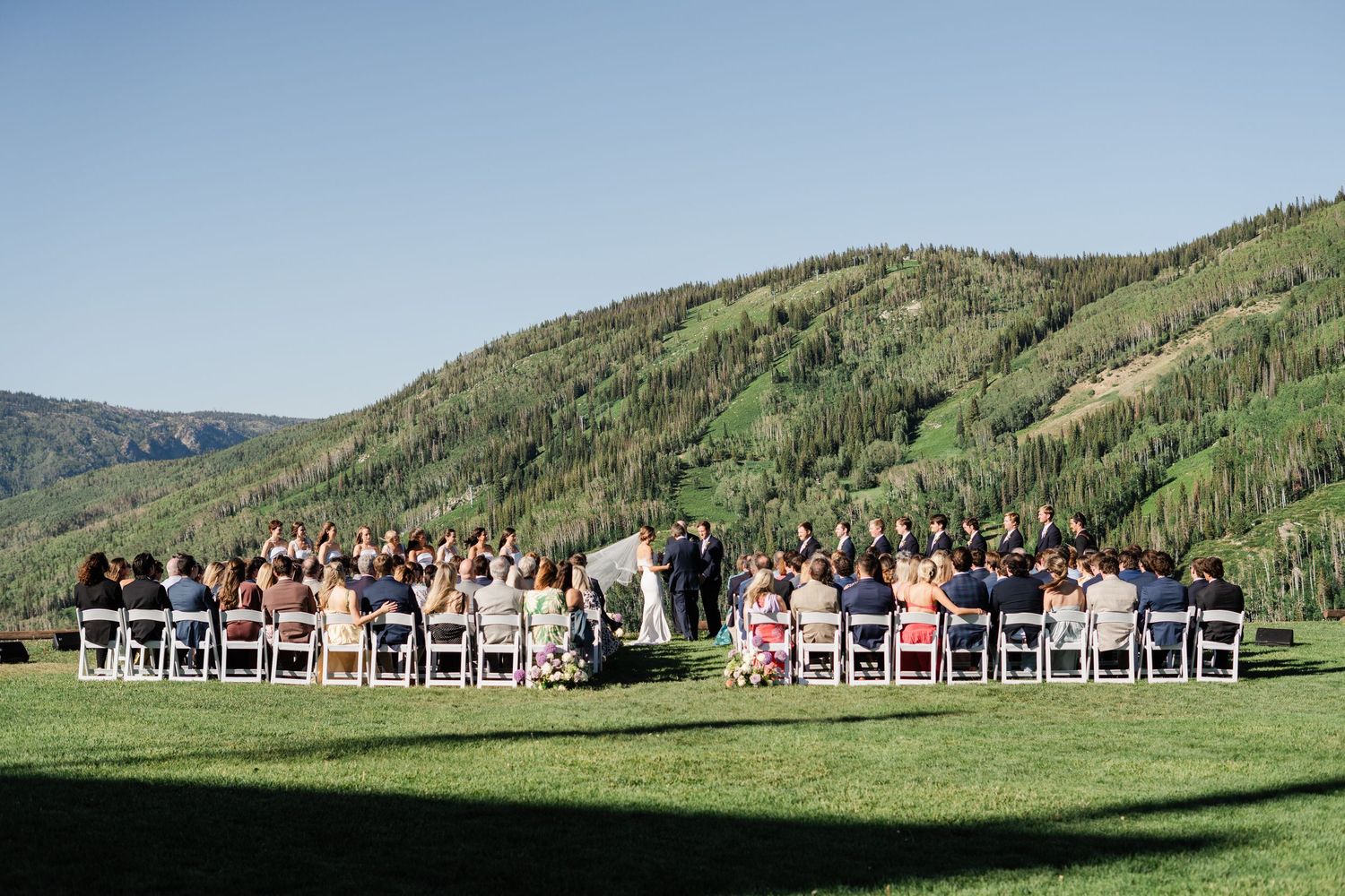 Outdoor mountain wedding ceremony with guests seated on white chairs on a green lawn.