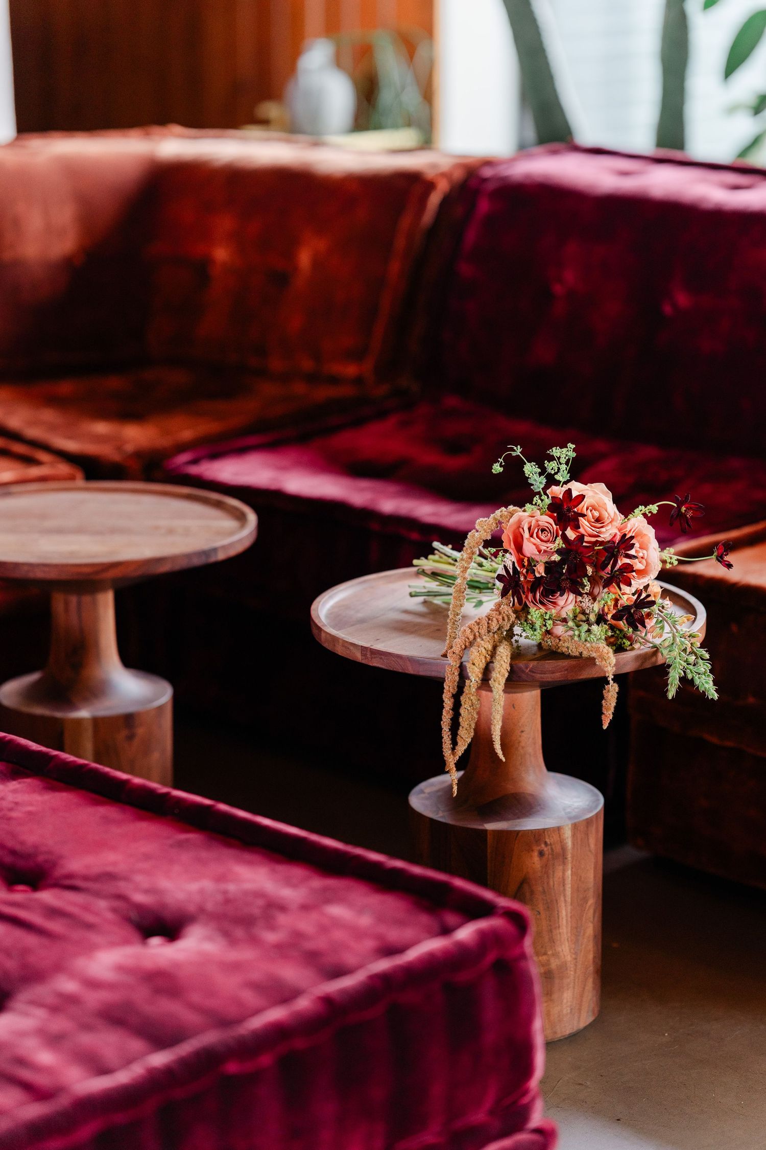 A rustic floral centerpiece with coral roses sits on a wooden pedestal beside plush burgundy velvet seating.
