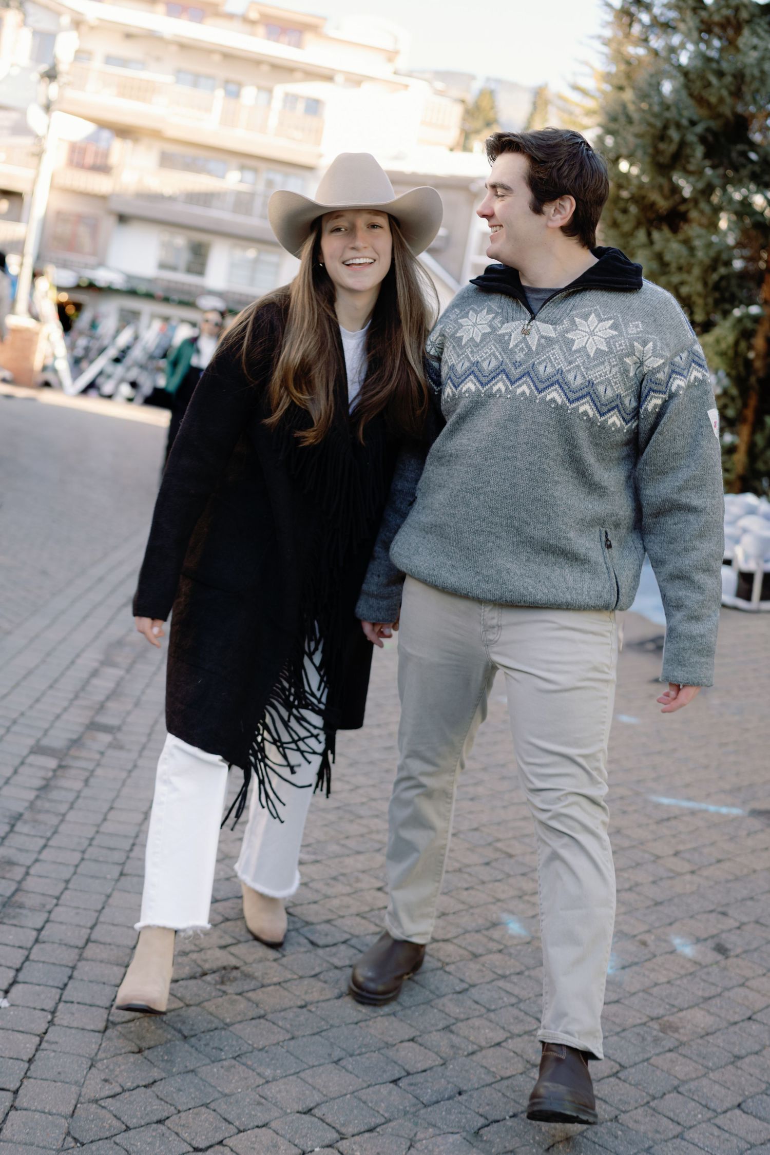 Stylish couple in winter outfits walking hand in hand through a snowy mountain village.