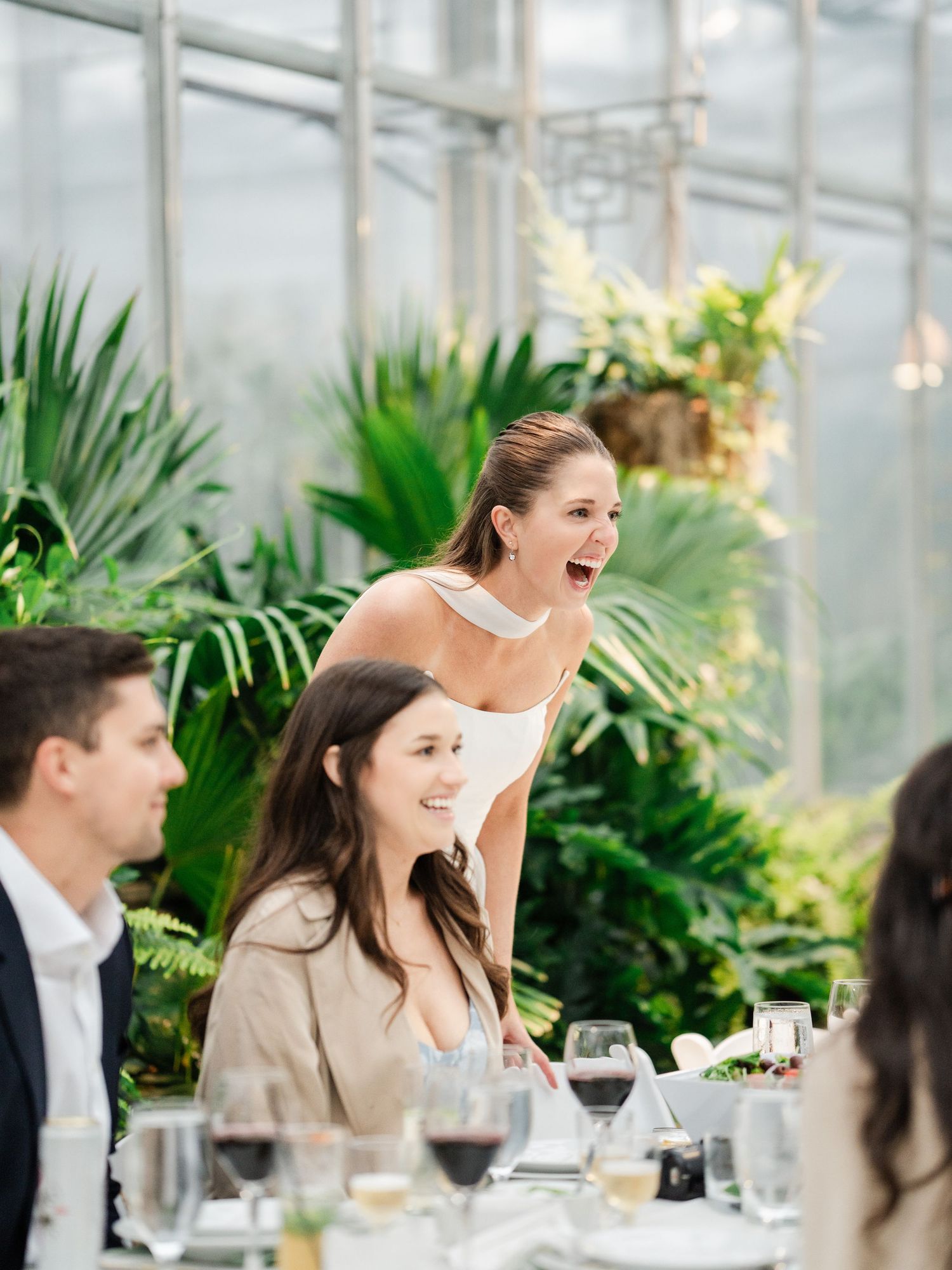 Joyful wedding guest laughing at reception table in lush greenhouse venue with tropical plants.