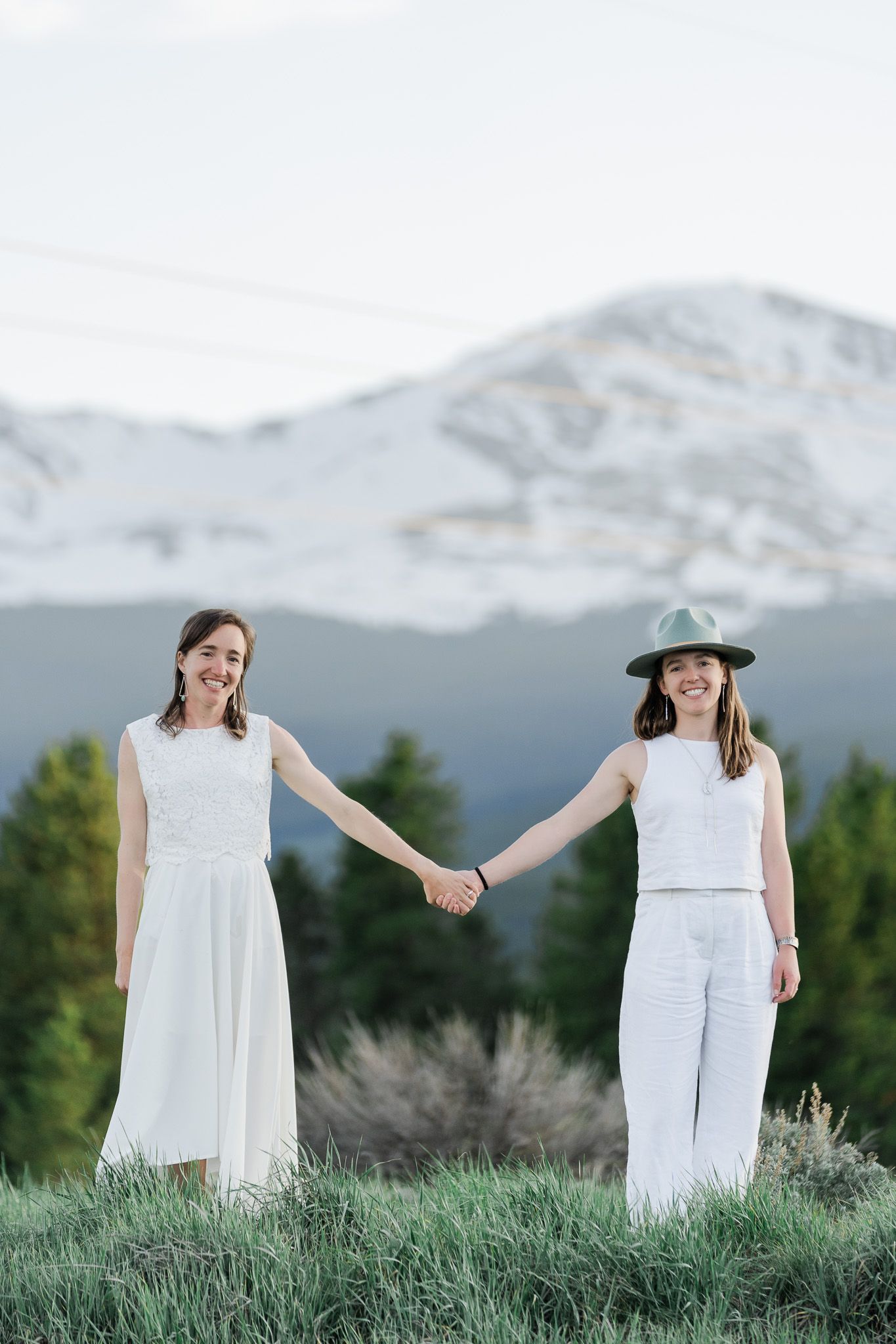 Two brides in white holding hands in a mountain meadow with snow-capped peaks and pine trees behind.