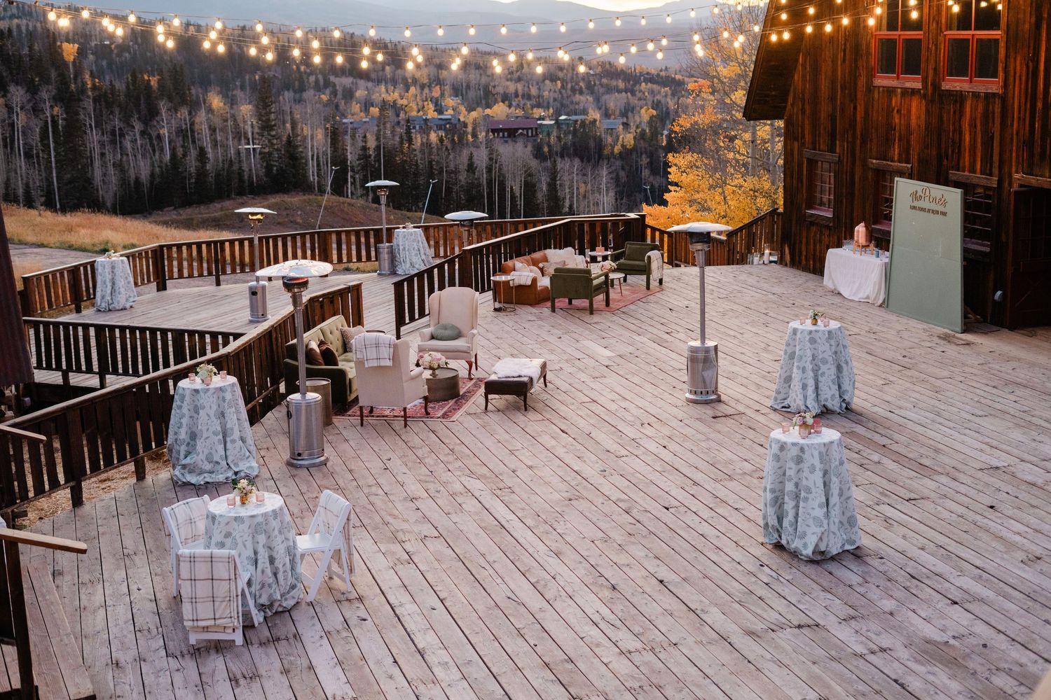 String-lit outdoor wedding reception deck with lounge seating and mountain forest backdrop at dusk.