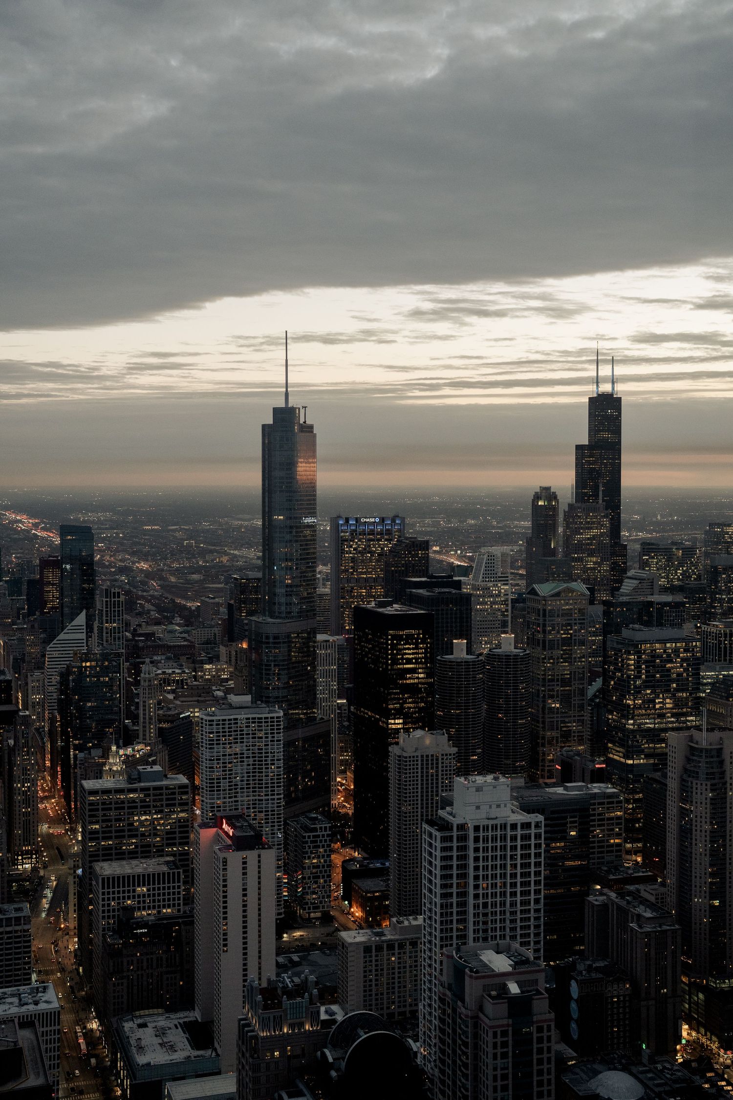 Dramatic aerial dusk view of Chicago skyline with Willis Tower and illuminated skyscrapers under cloudy sky.