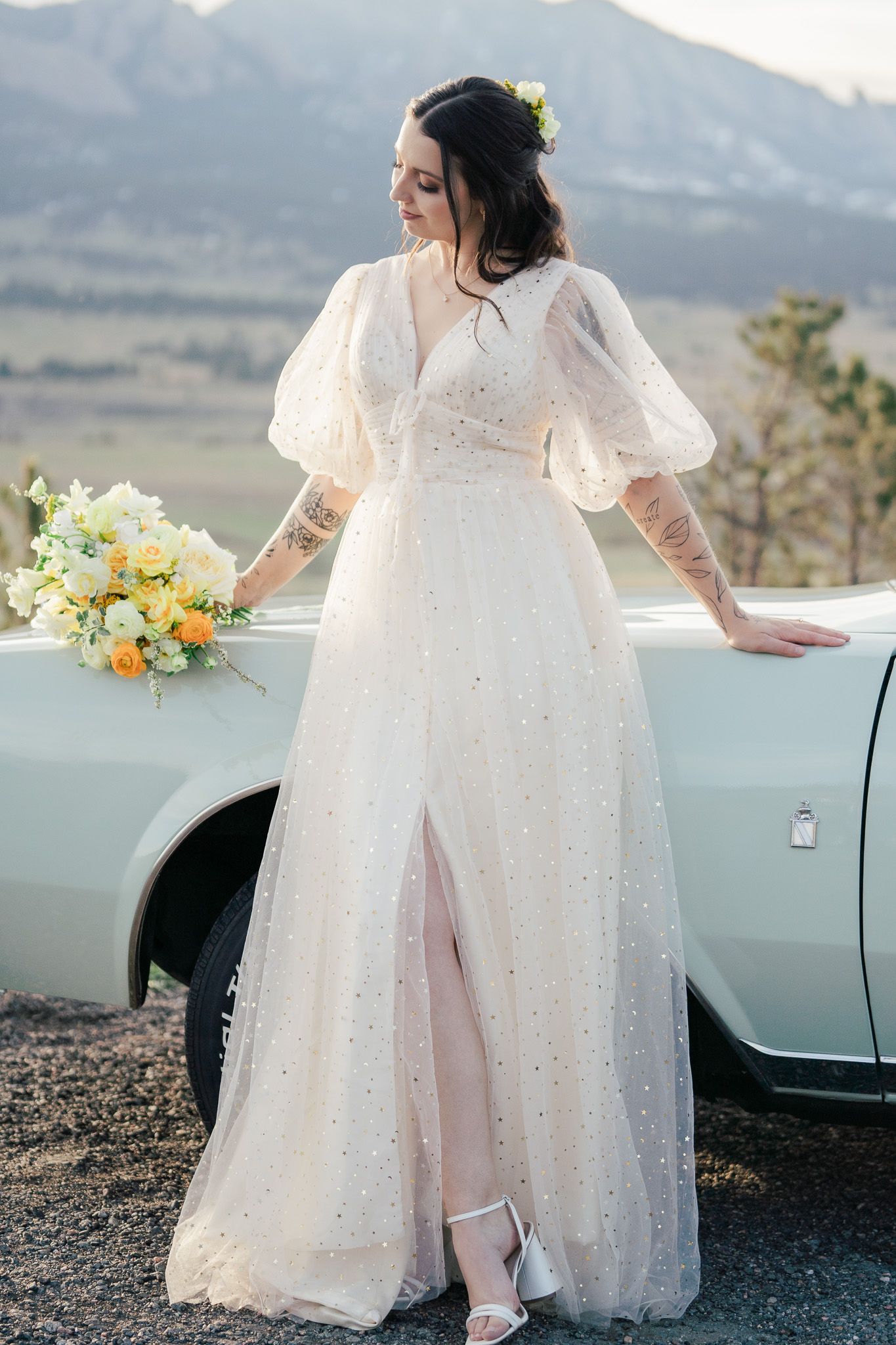 Bride in pearl-dotted tulle gown with puff sleeves holding yellow bouquet beside vintage white car outdoors.