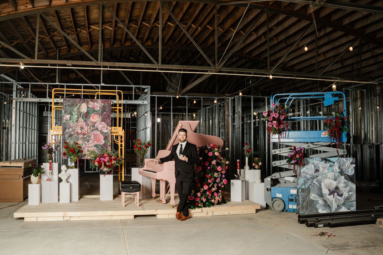 Man in suit stands beside a pink piano surrounded by floral arrangements in an industrial venue with metal scaffolding.