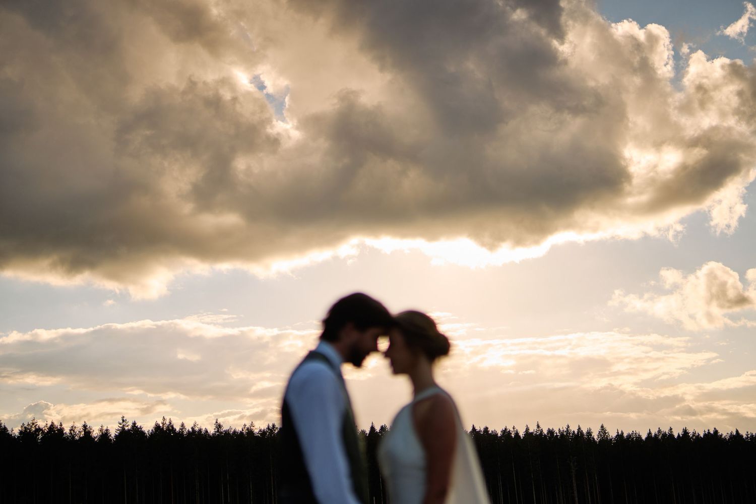 Silhouetted figures stand against a dramatic sunset sky with glowing clouds and a forest treeline in the background.