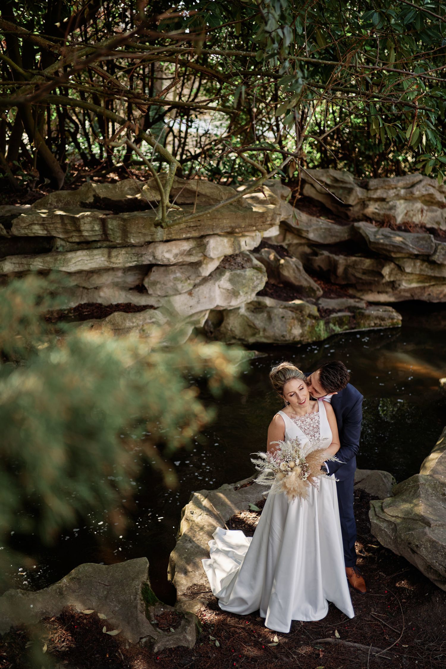 A romantic couple embraces by a serene pond surrounded by natural rock formations and lush greenery.