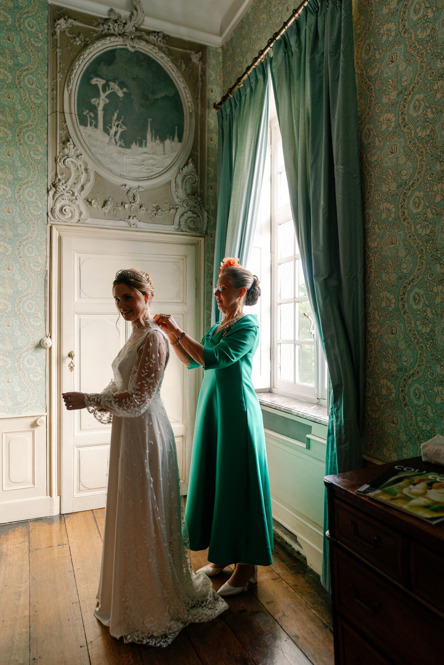 A figure in a vintage white dress and green coat stands near ornate walls and emerald curtains in a historic room.