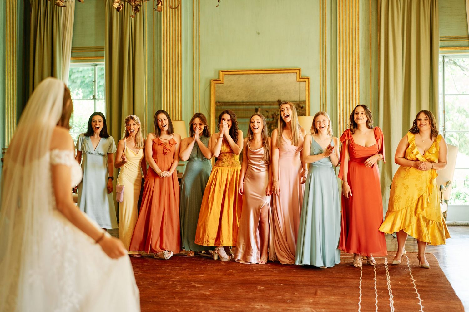 A wedding party in an elegant room wearing colorful bridesmaid dresses in shades of coral, orange, pink and blue.