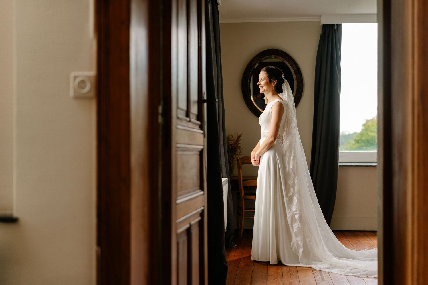 Bride in flowing white wedding dress stands elegantly in a sunlit hallway with dark wood accents and mirrors.
