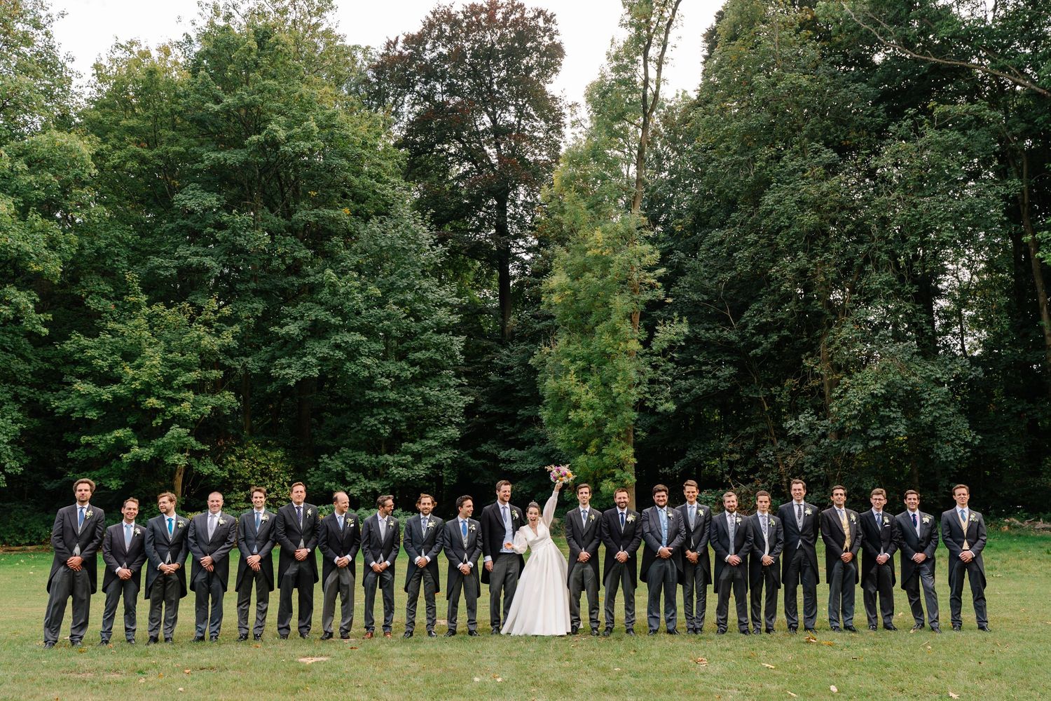 Wedding party lined up outdoors in formal attire against lush green trees in a garden setting.