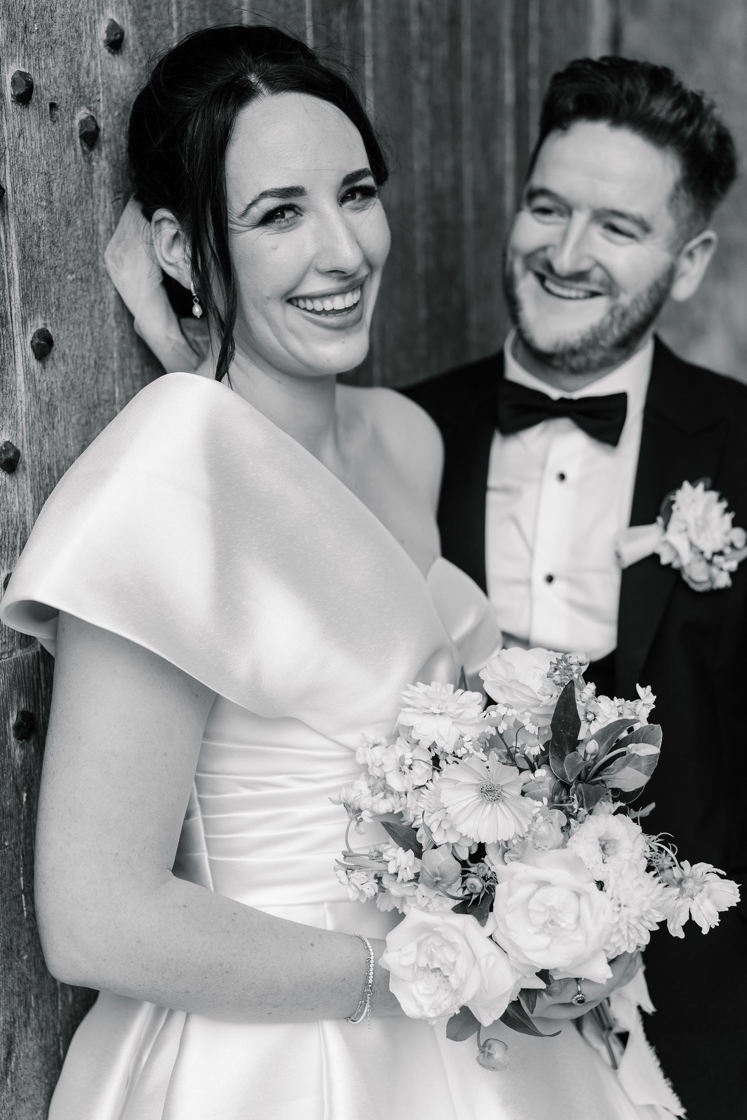 Black and white wedding portrait of a couple smiling with the bride holding a white floral bouquet.