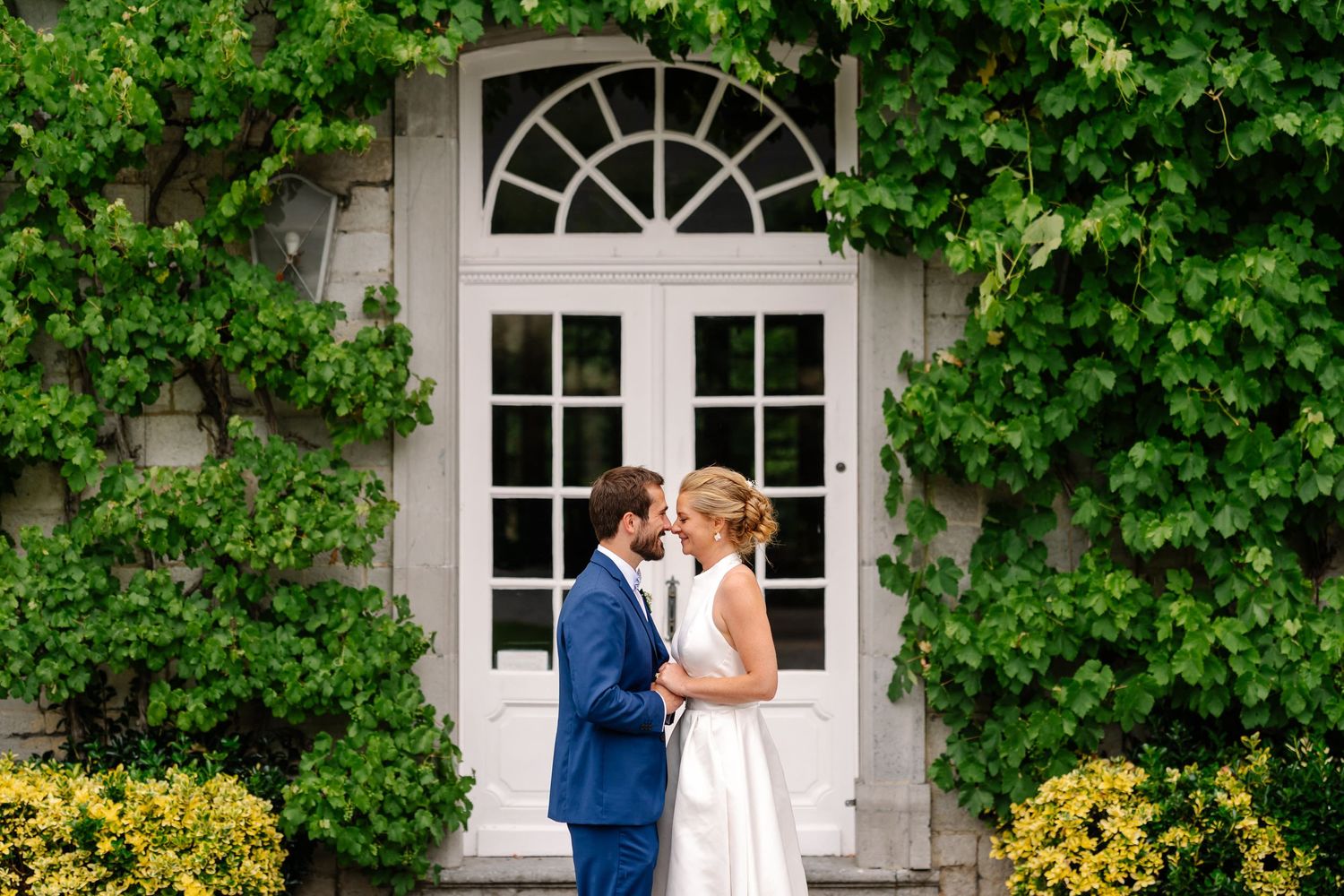 A couple shares a romantic moment in front of a white arched doorway surrounded by lush green vines and yellow flowers.